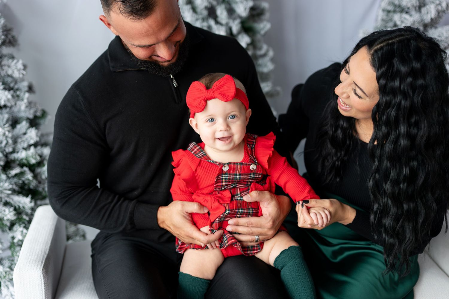Happy baby girl in red plaid dress smiles during Christmas family photo session with parents on white bench.
