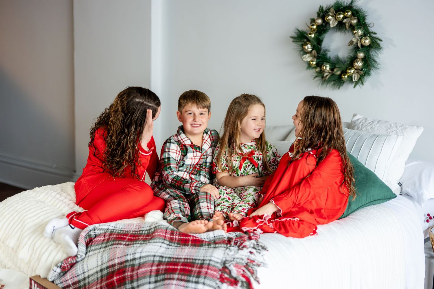 Four children in festive Christmas pajamas sit together laughing on a bed decorated for the holidays.
