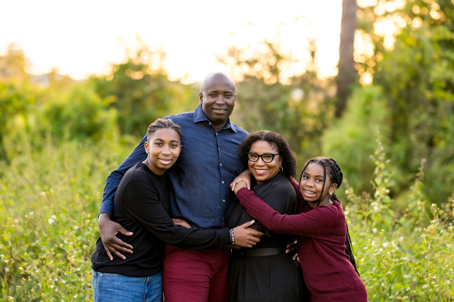 A family embraces while standing together in a sunlit meadow with wildflowers during golden hour.