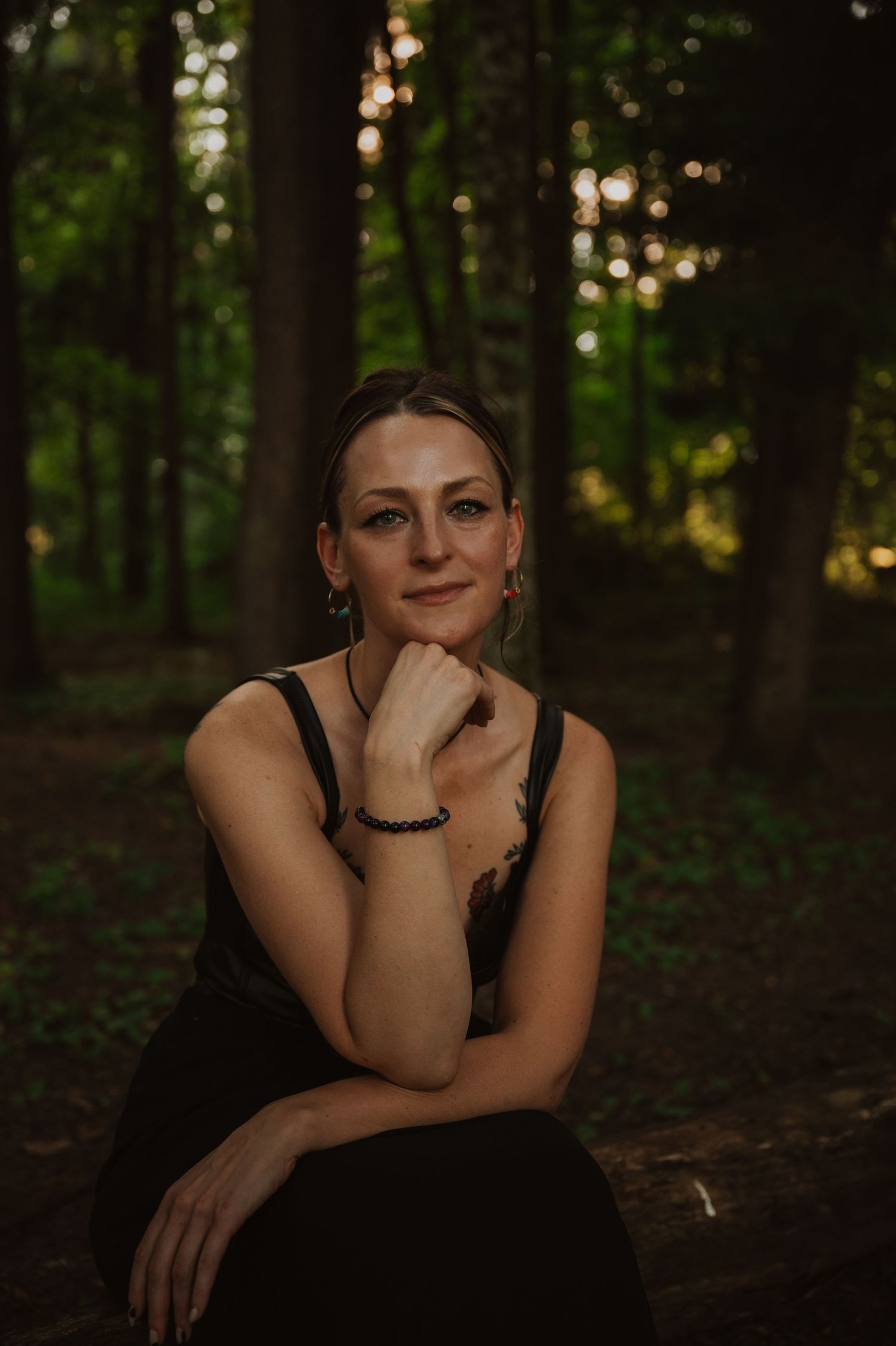 Portrait in a black dress sits thoughtfully in a dark forest setting with glowing bokeh lights in the background.