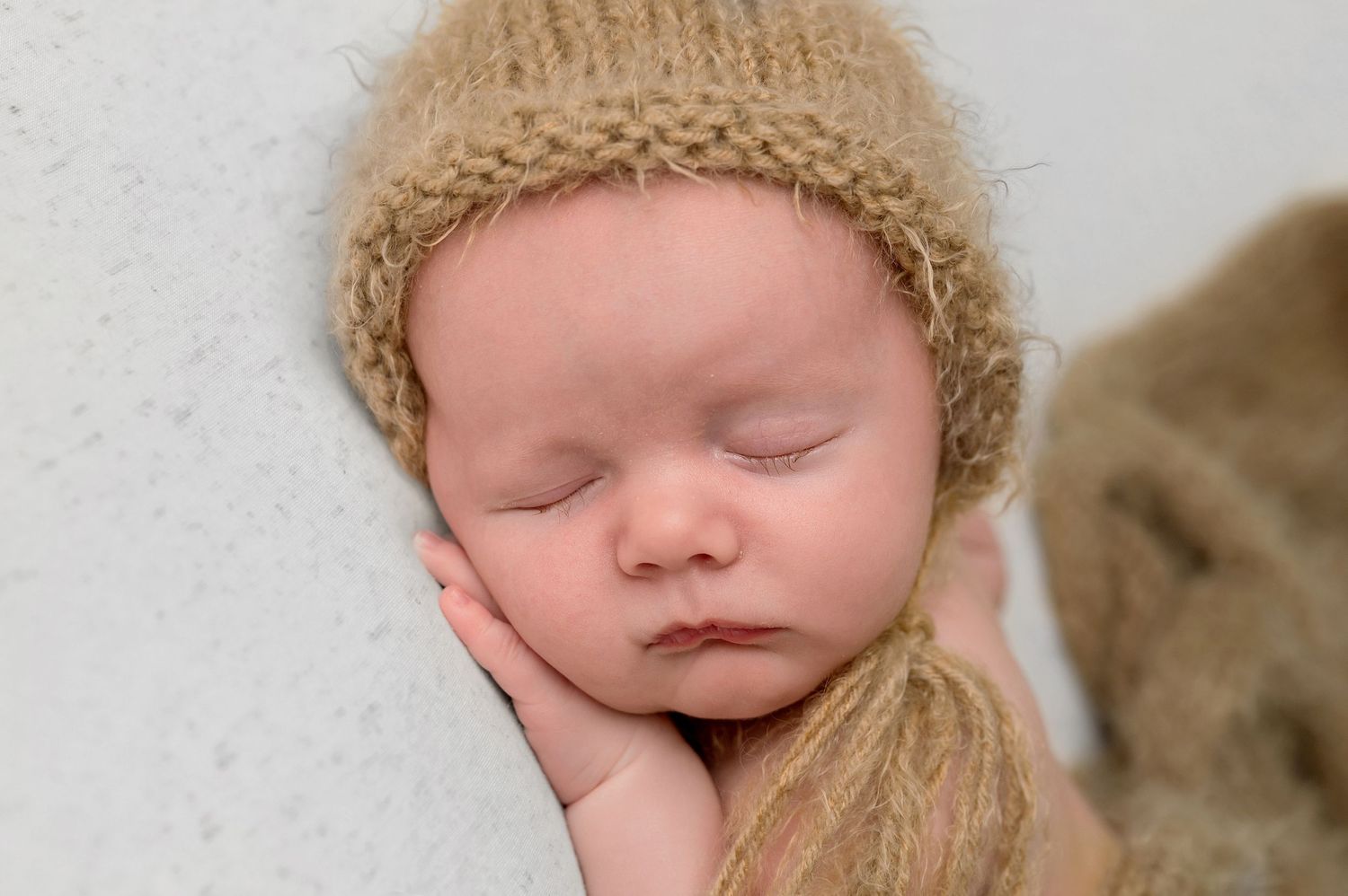 Sleeping newborn baby wearing a cozy knit hat rests peacefully against a soft beige backdrop.