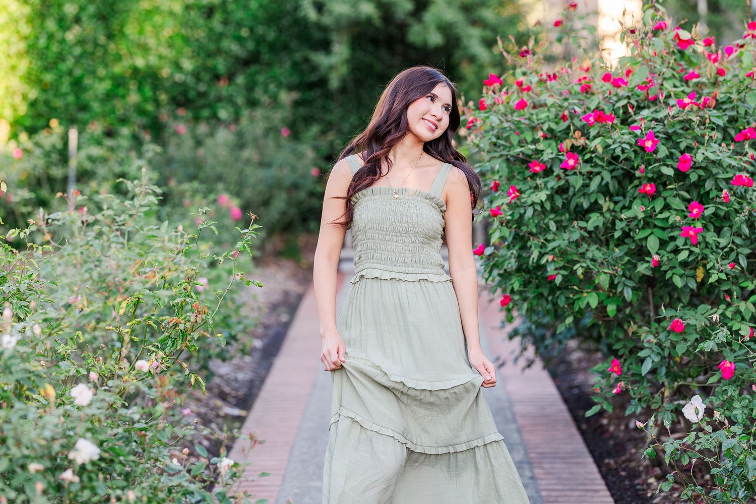 A person in a flowing sage green dress poses in a garden path surrounded by blooming pink rose bushes and greenery.