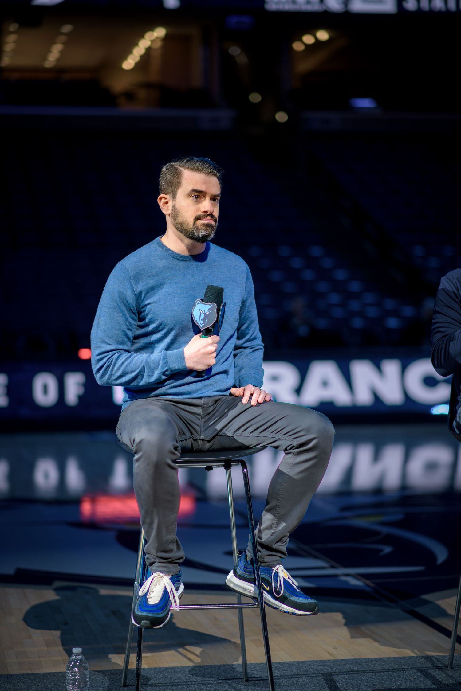 Basketball player stretches on chair before game wearing blue sweatshirt and grey pants in dark arena.