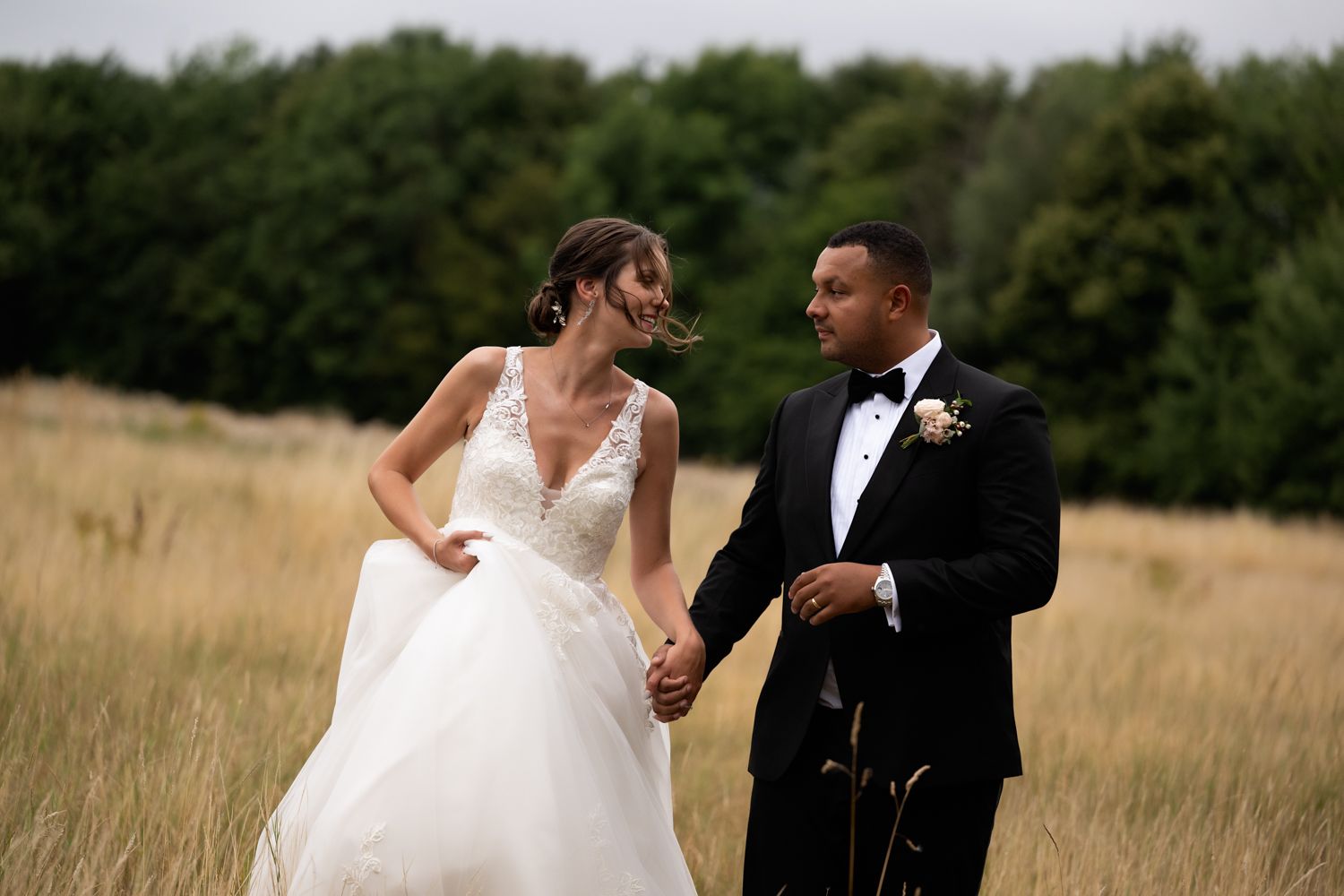 A couple in wedding attire holds hands while walking through a golden field with trees in the background.