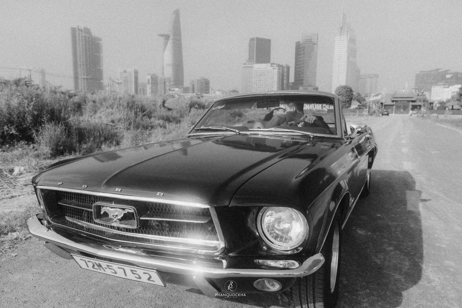 Black and white photo of classic Ford Mustang parked with Ho Chi Minh City skyline in background.