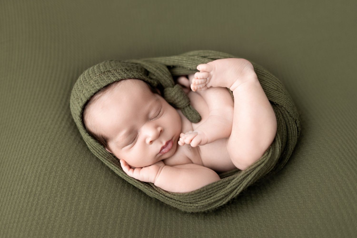Newborn baby sleeping peacefully wrapped in olive green knit blanket in professional photography studio setting.