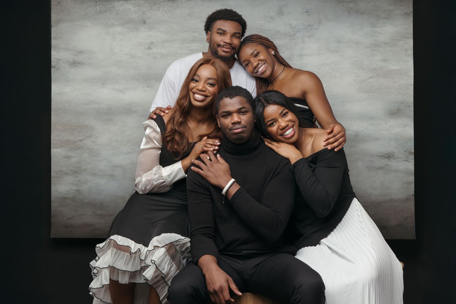 Family of five posing in a studio portrait wearing coordinated black, cream, and neutral outfits against a textured gray backdrop.