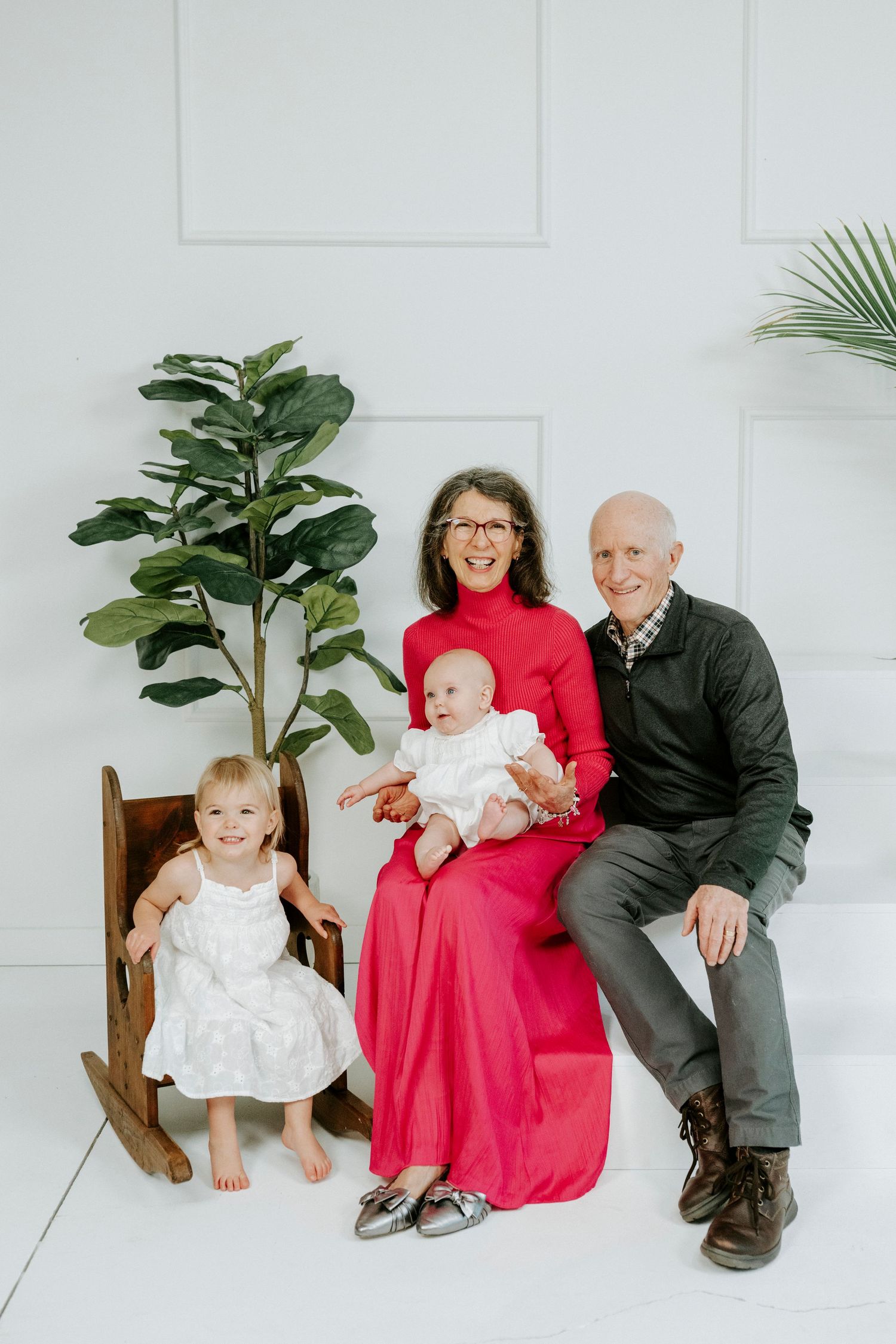 A family photo sequence showing a baby, woman in red dress, and man in black suit sitting against a white wall with plants.