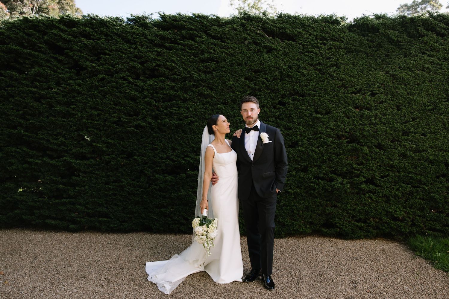 A couple poses in front of a lush green hedge during an elegant outdoor wedding ceremony.