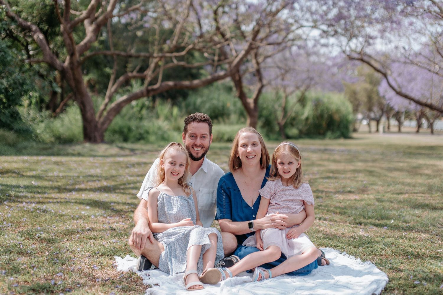 Family sitting together on white blanket in grassy field during spring season with blooming trees in background.