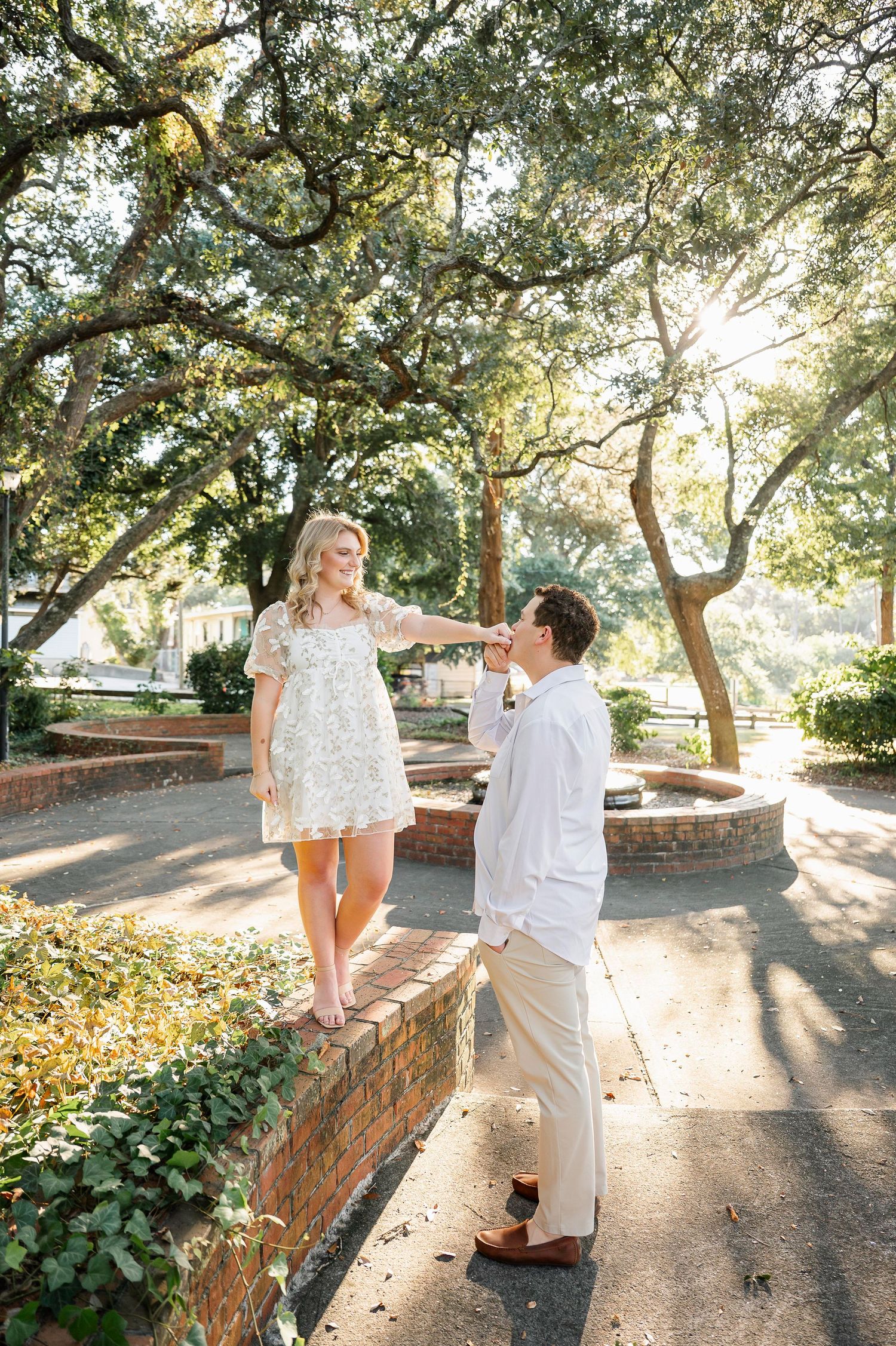 A couple shares playful moments along a sunlit brick wall under oak trees.