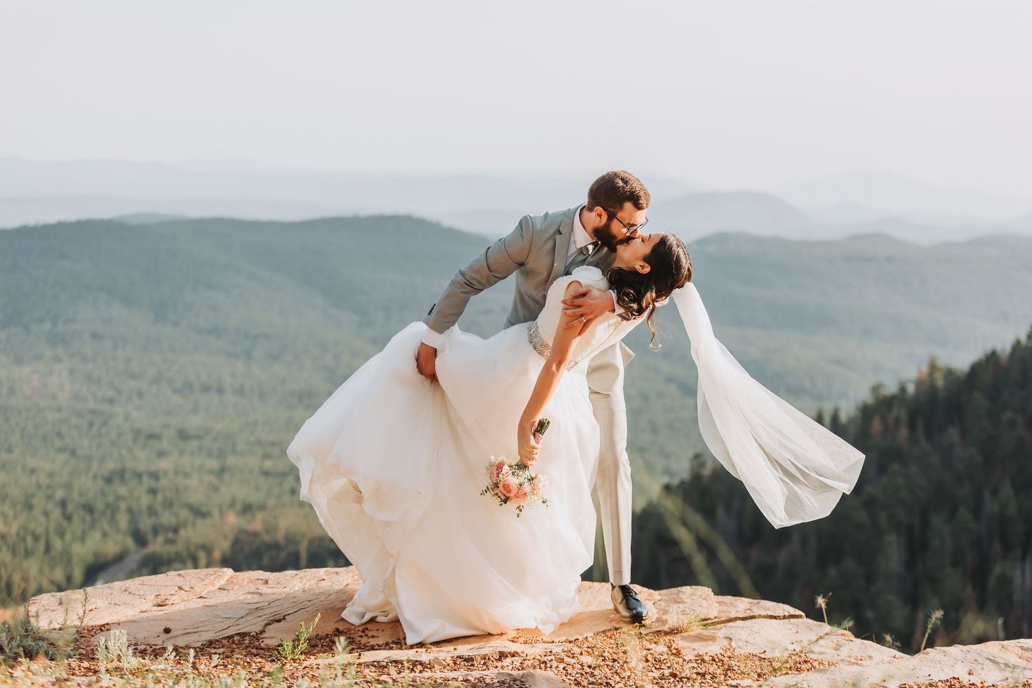 A couple in wedding attire embraces on a scenic mountain overlook as the bride's white dress billows in the wind.