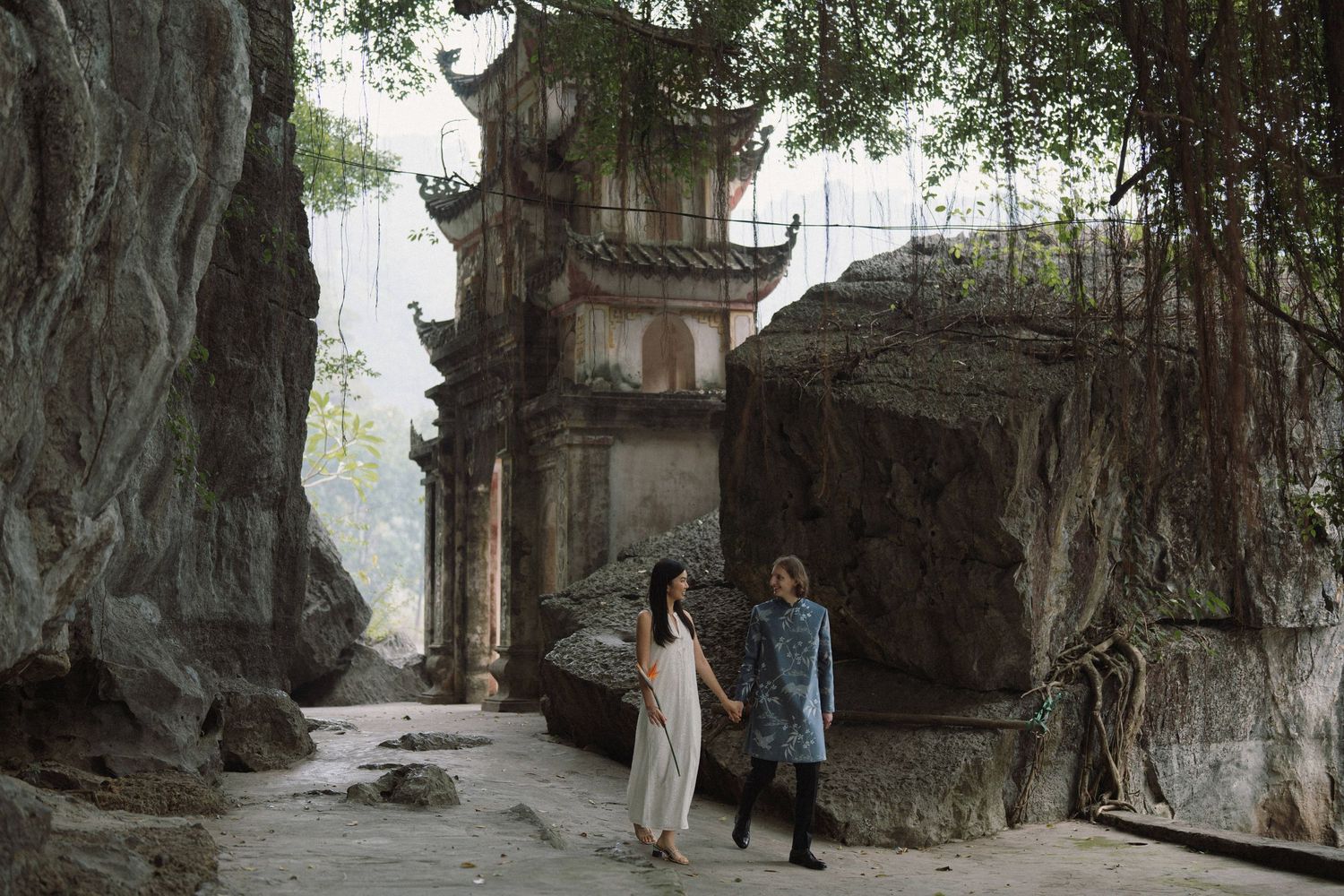 People walking alongside impressive banyan tree roots and stone walls in a historic temple or fortress setting.