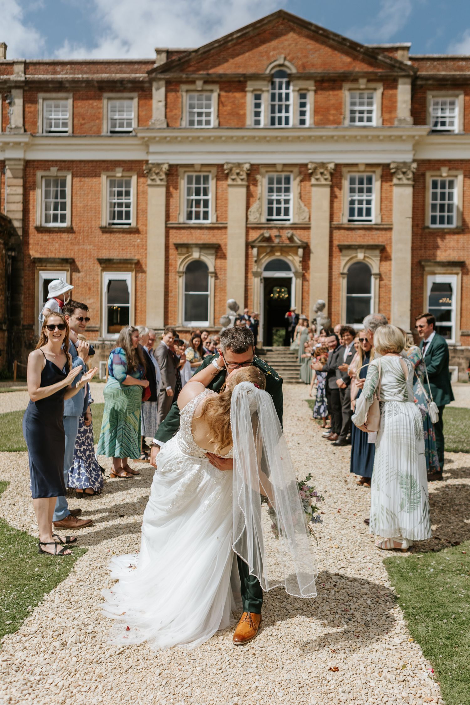 A newlywed couple walks down a tree-lined path together, with the bride in white and groom in emerald green.