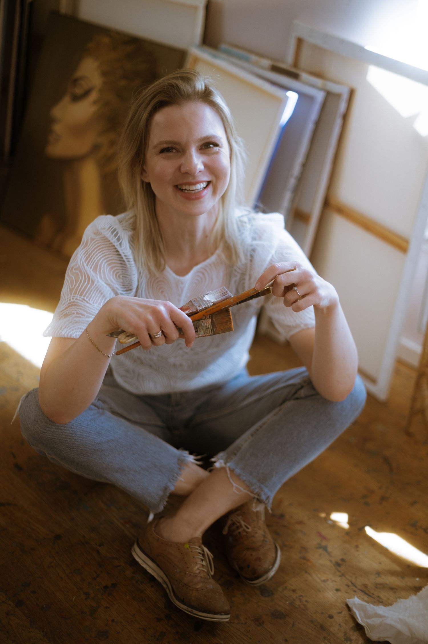 Artist sits cross-legged on wooden floor holding art supplies in casual attire.