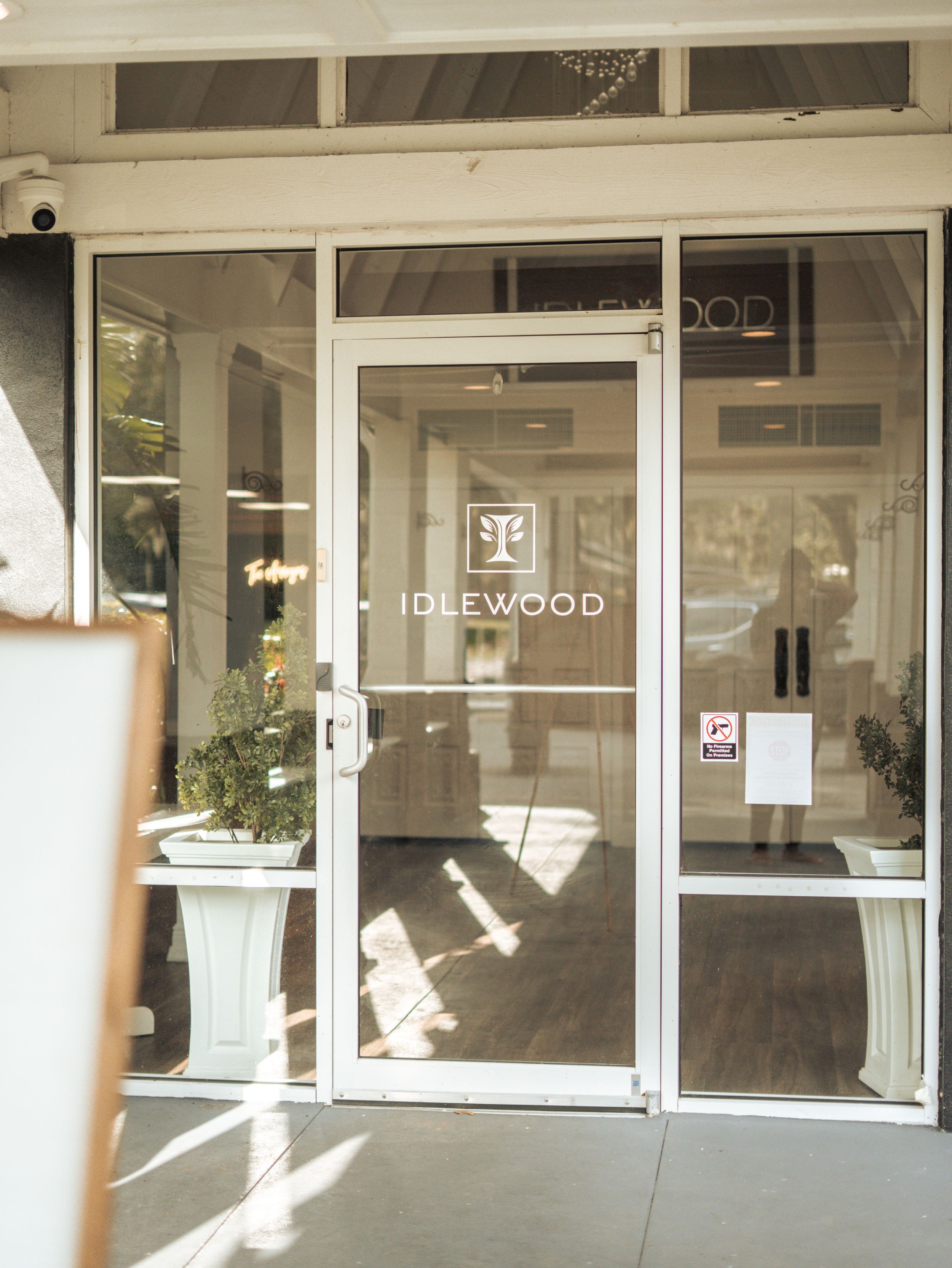 Glass storefront entrance of Idlewood building with white lettering and sunlight streaming through the glass door.