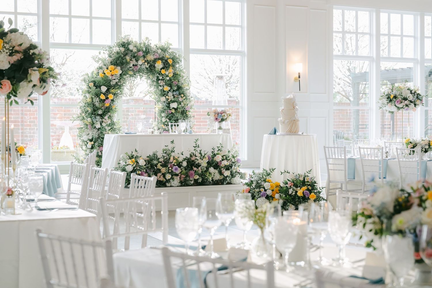 Elegant wedding reception venue with white chairs, floral arches, and natural light streaming through large windows. - The Crawford