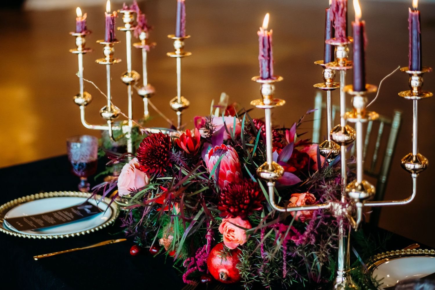 Elegant table setting with gold candlesticks and deep red floral arrangements on a black tablecloth.