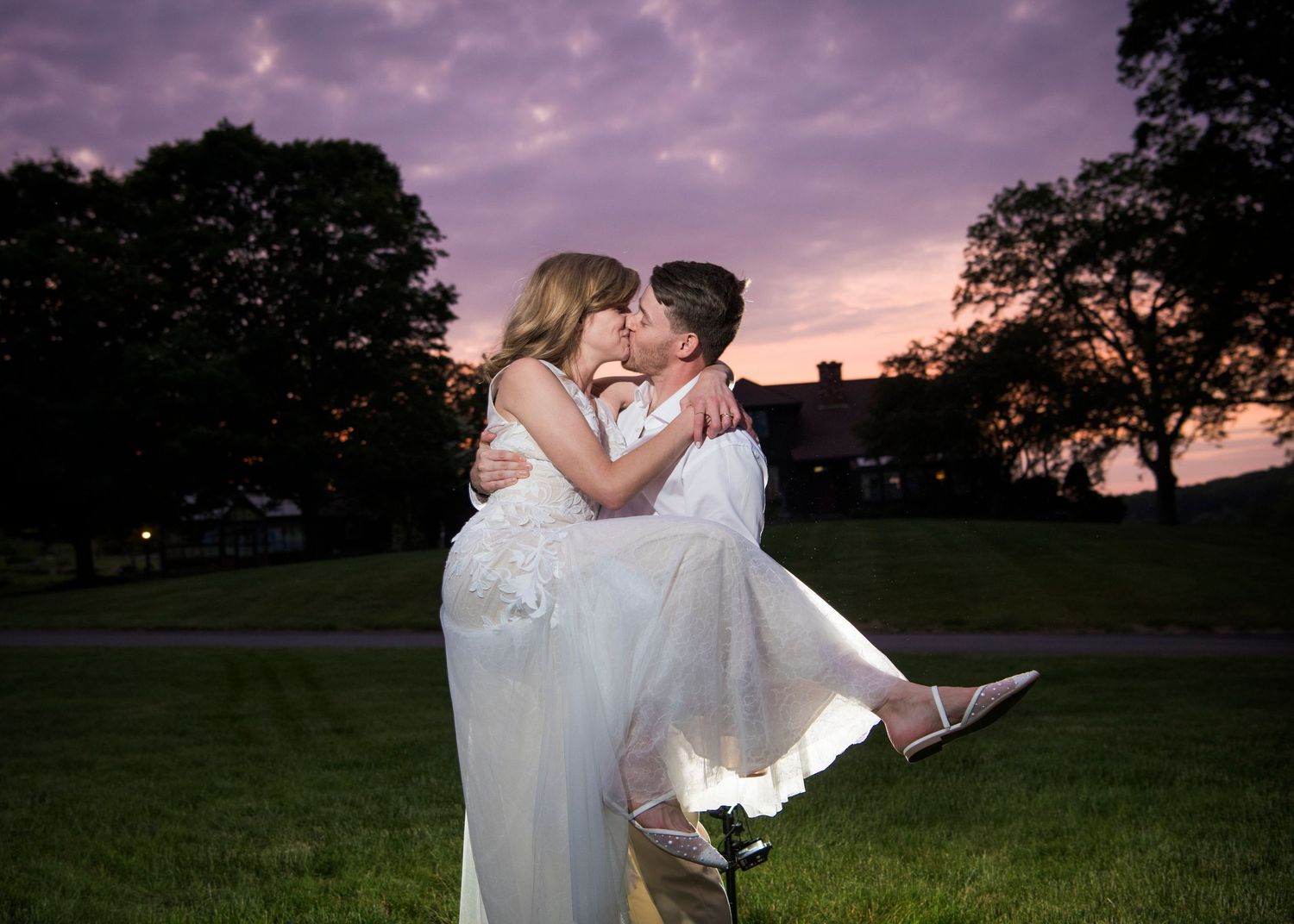 Newlyweds share an intimate moment outdoors with autumn foliage and rustic architecture in background.