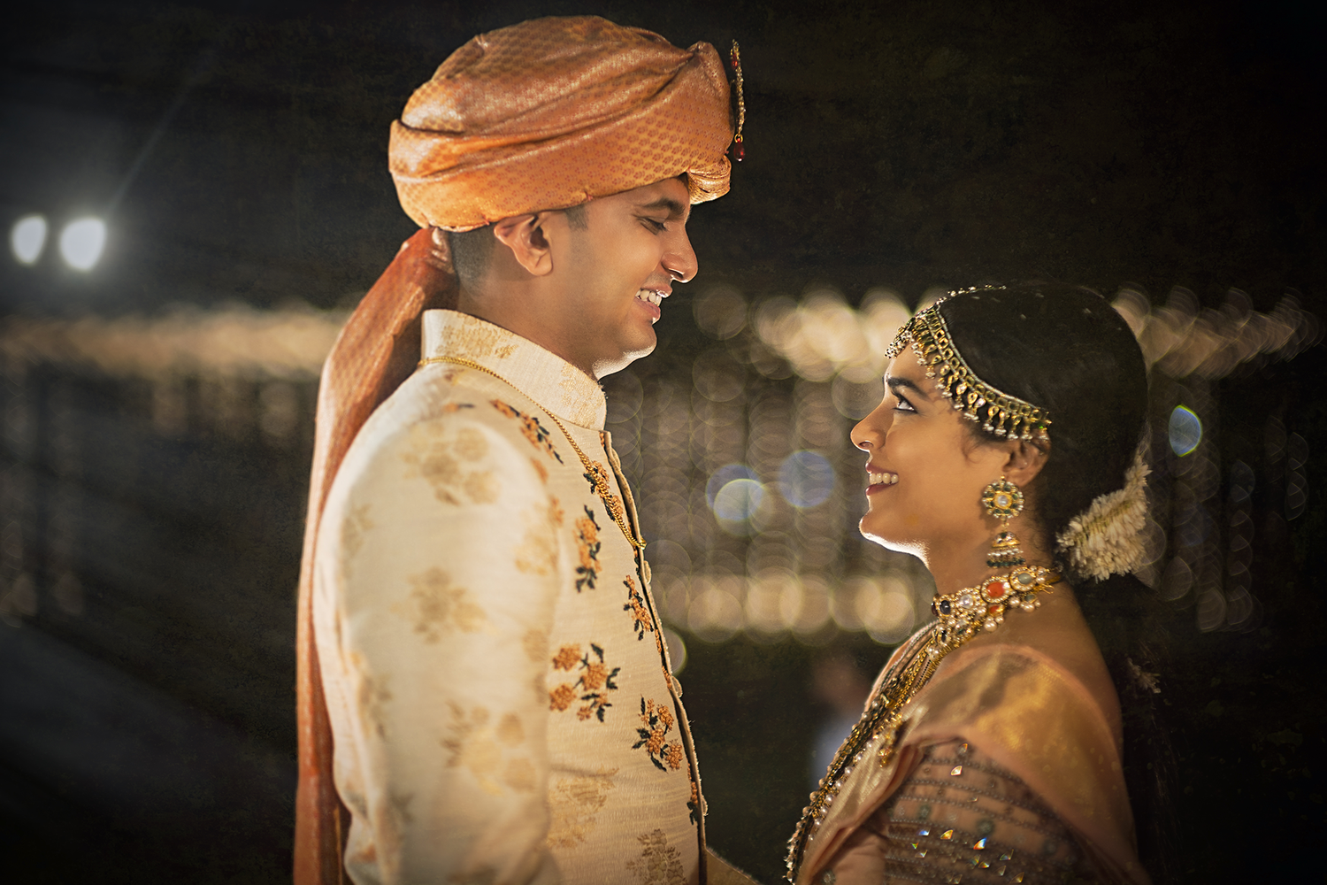 A Indian couple stands intimately in traditional Indian wedding attire with ornate gold details and jewelry in a low-lit setting.