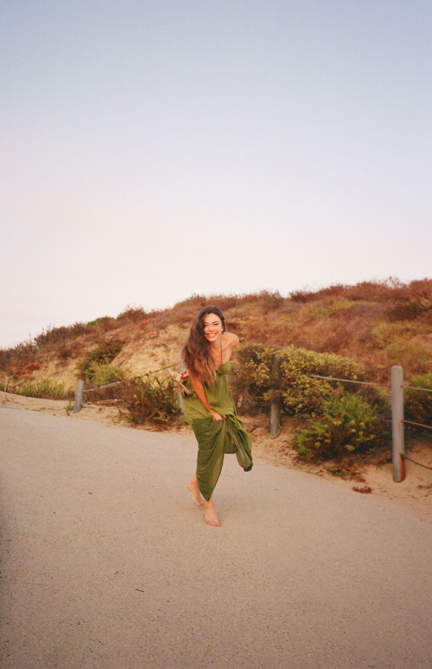 Woman in flowing green dress dances on dirt path near coastal hillside at sunset.