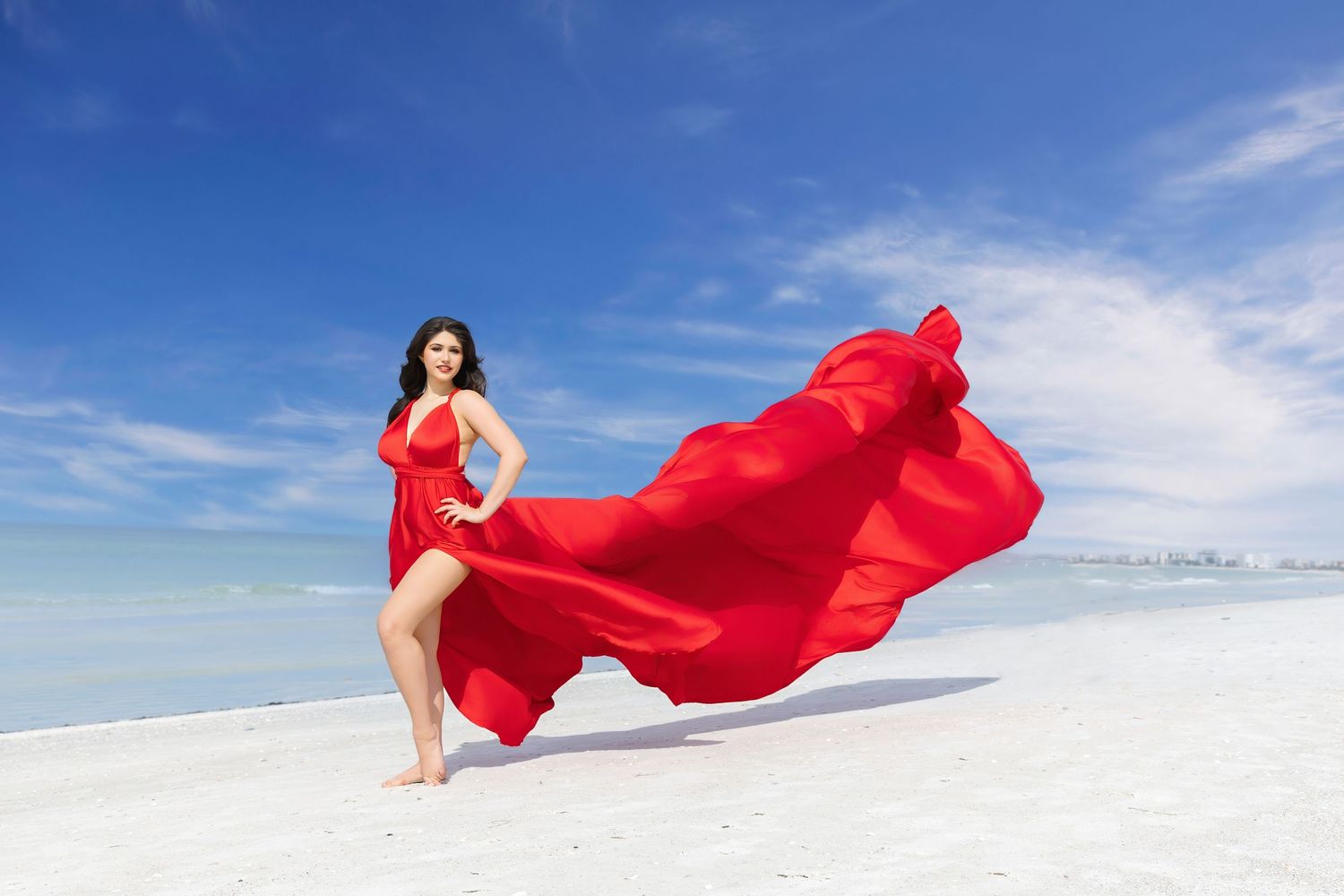 Flowing red fabric dress billows dramatically against white sand and blue sky on a tropical beach. Flying Dress Photoshoot
