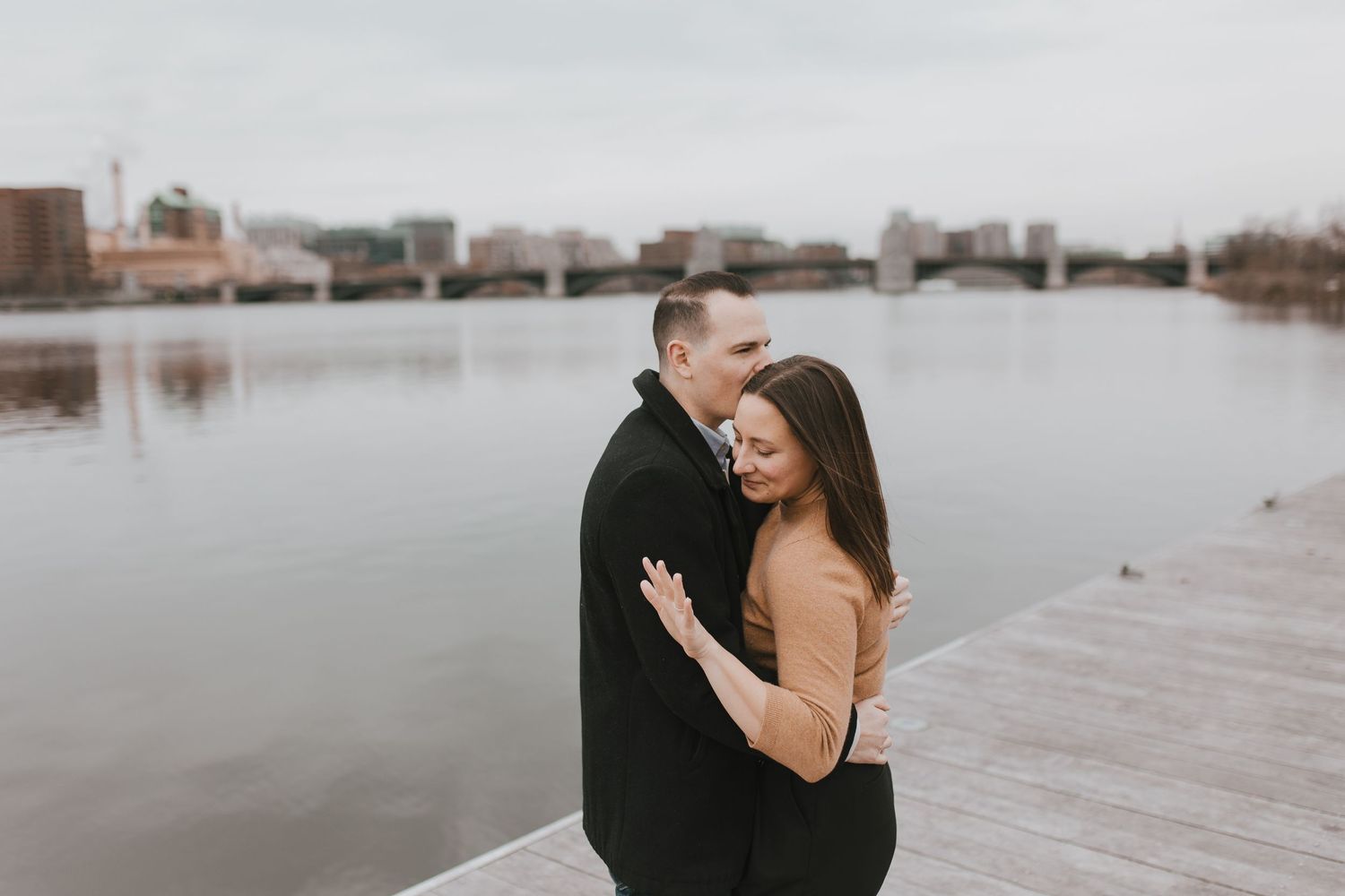 A couple shares an intimate moment on a waterfront dock with city buildings visible in the distance.