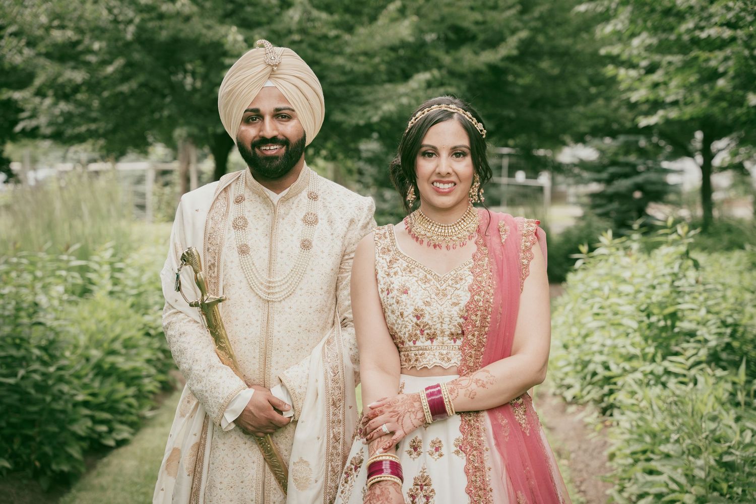 Traditional Sikh wedding couple in ornate cream and pink wedding attire pose in garden setting.