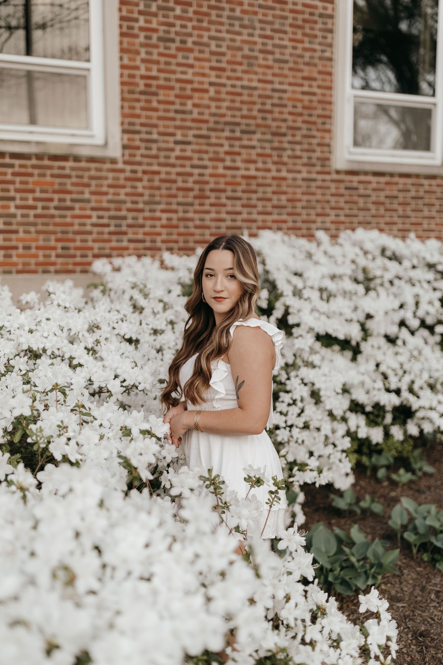 Graduate in white dress poses among blooming white azalea bushes near brick building on college campus.