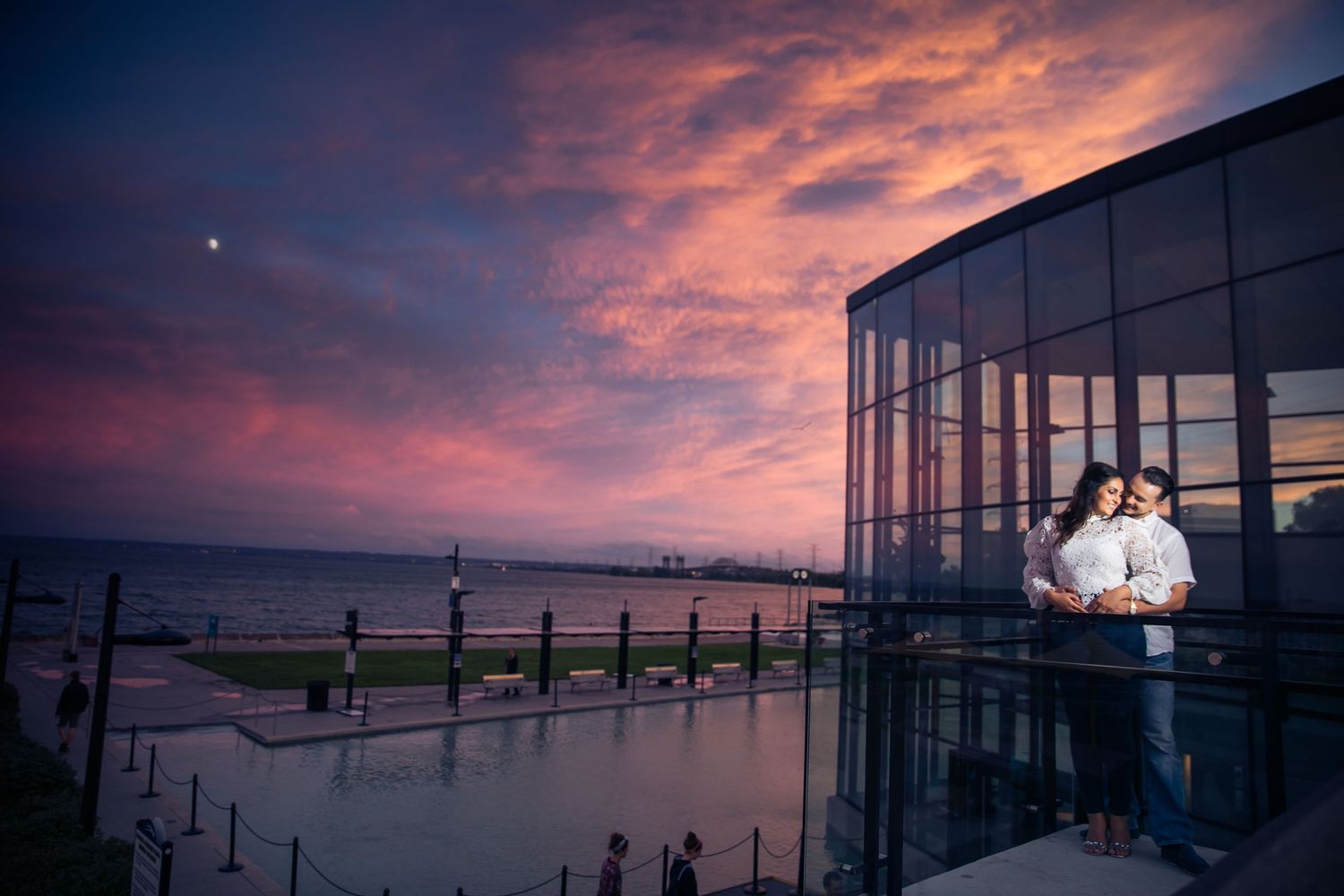 Couple silhouetted against dramatic sunset sky at modern glass venue overlooking waterfront.