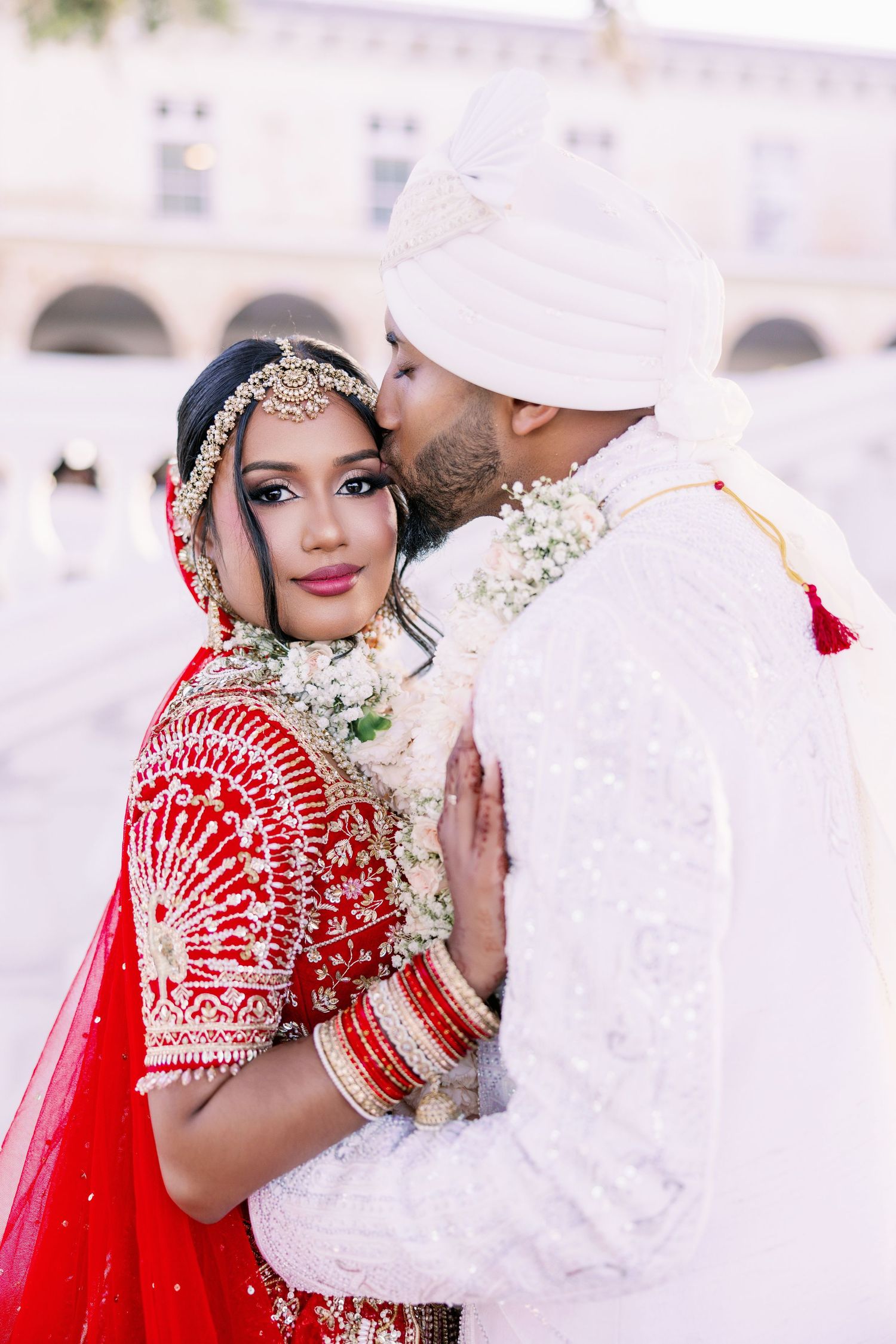 Traditional Indian wedding moment with newlyweds in white and red attire embracing against an elegant arched backdrop.
