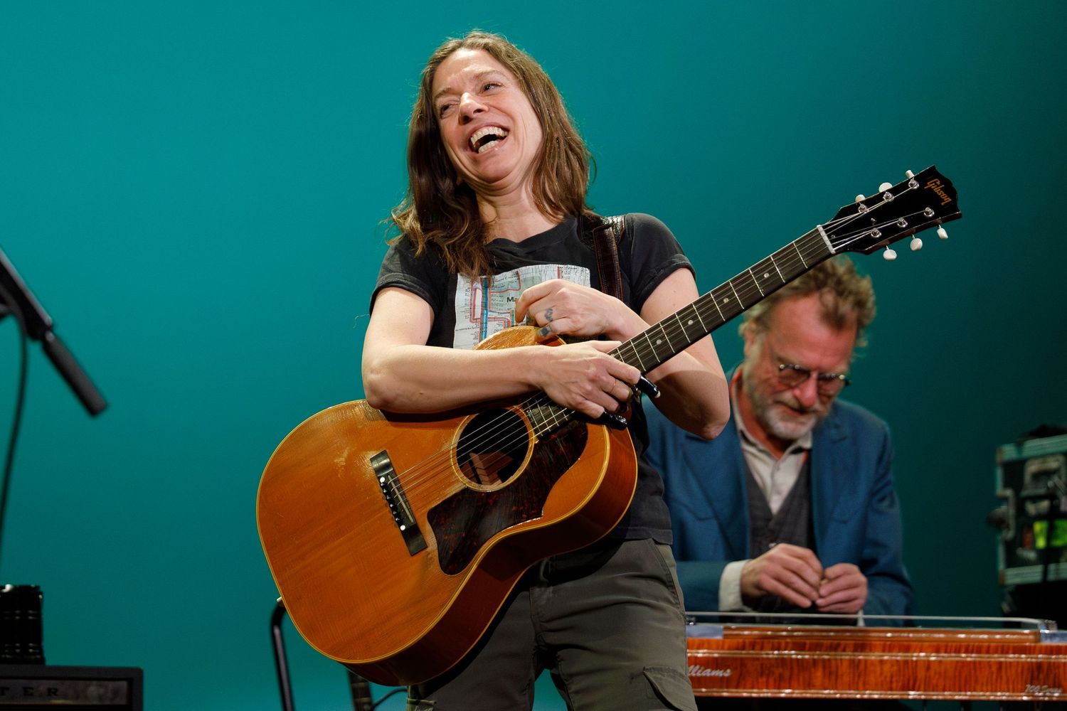Musician playing acoustic guitar on stage with joyful expression while performer with lap steel guitar sits in background.