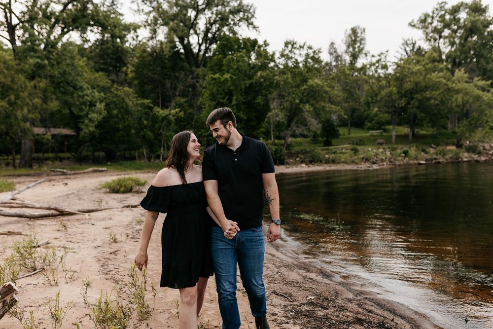 A couple wearing casual black outfits strolls along a peaceful riverside beach surrounded by green trees.