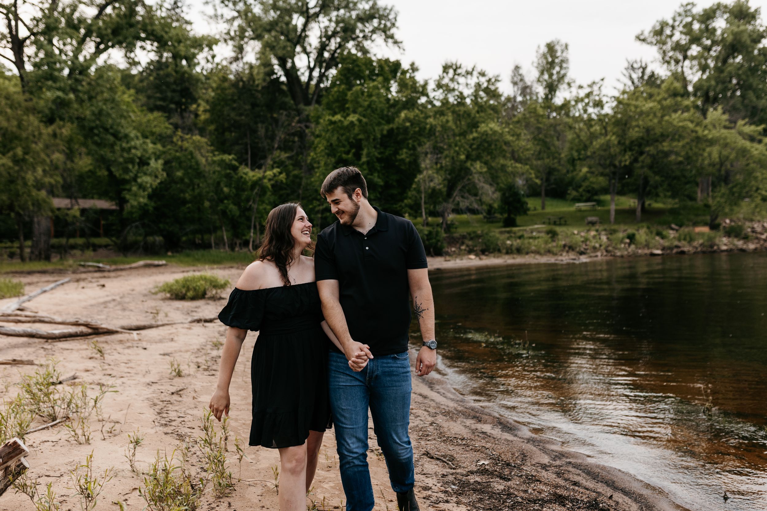A couple wearing casual black outfits strolls along a peaceful riverside beach surrounded by green trees.