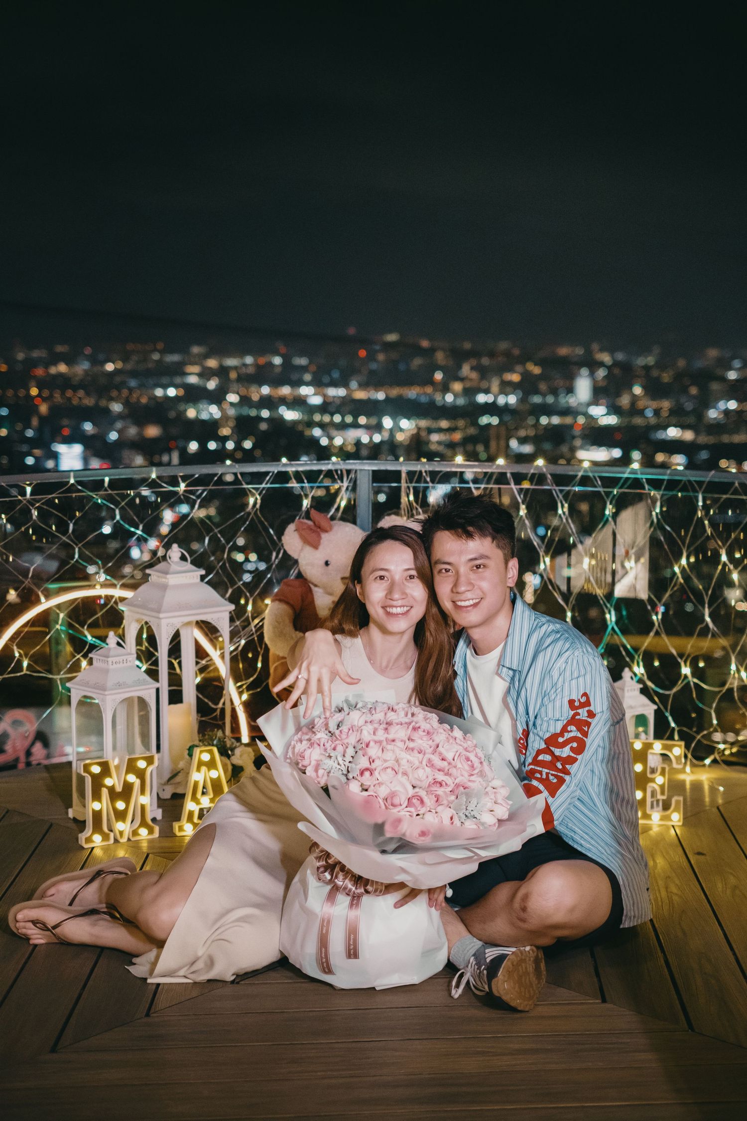 Happy couple sitting on rooftop at night holding large pink flower bouquet surrounded by fairy lights.