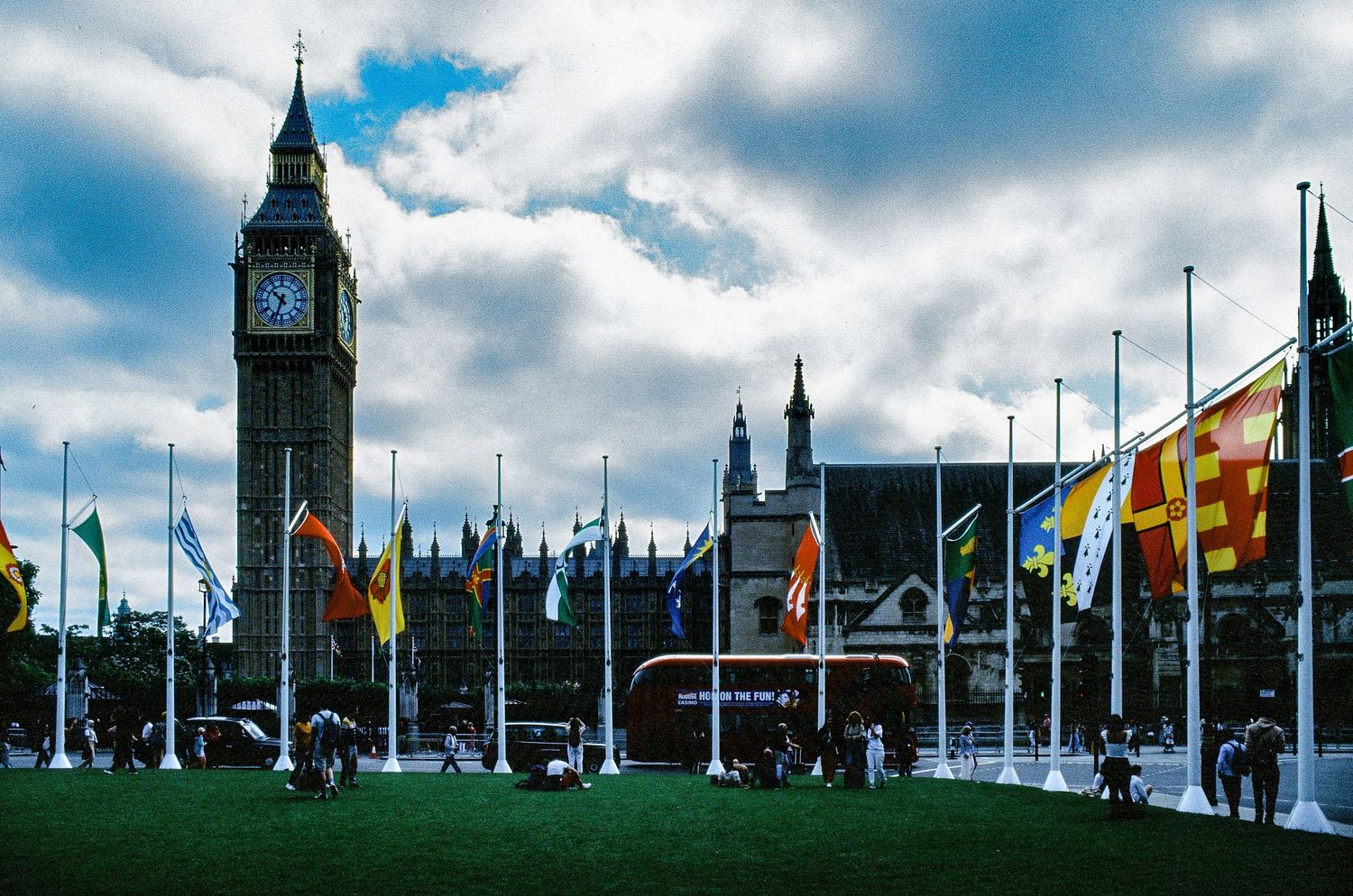 Big Ben and the Houses of Parliament in London with colorful flags and cloudy blue sky.