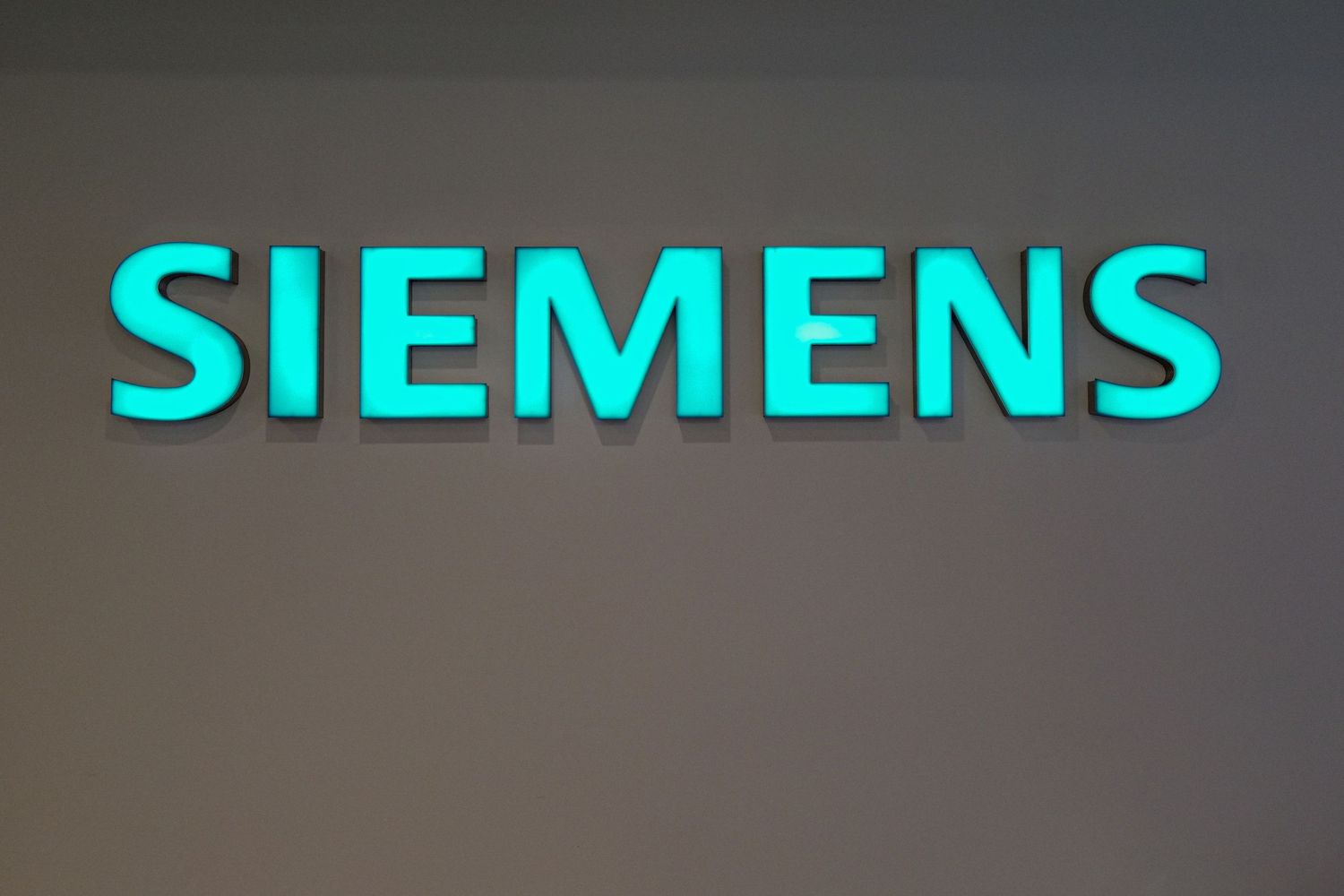 Illuminated teal Siemens logo sign mounted on a gray wall in a corporate setting.