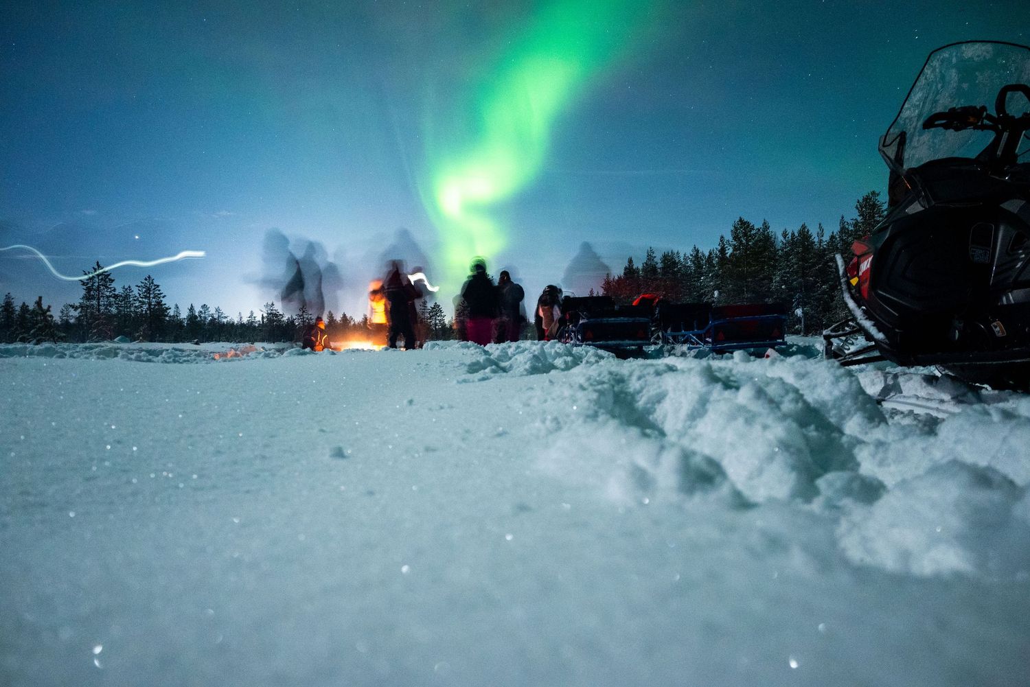 Northern lights illuminate a snowy landscape with snowmobiles in the foreground.