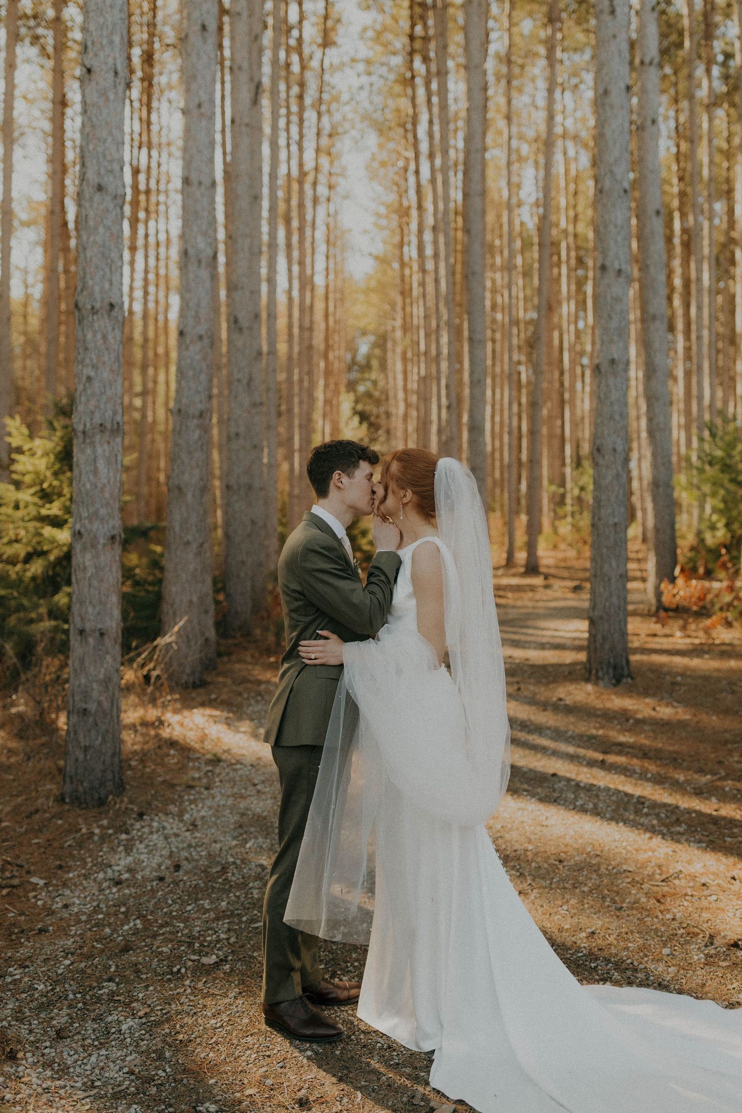 Wedding couple sharing intimate moment in sunlit pine forest with long white dress and fall colors.