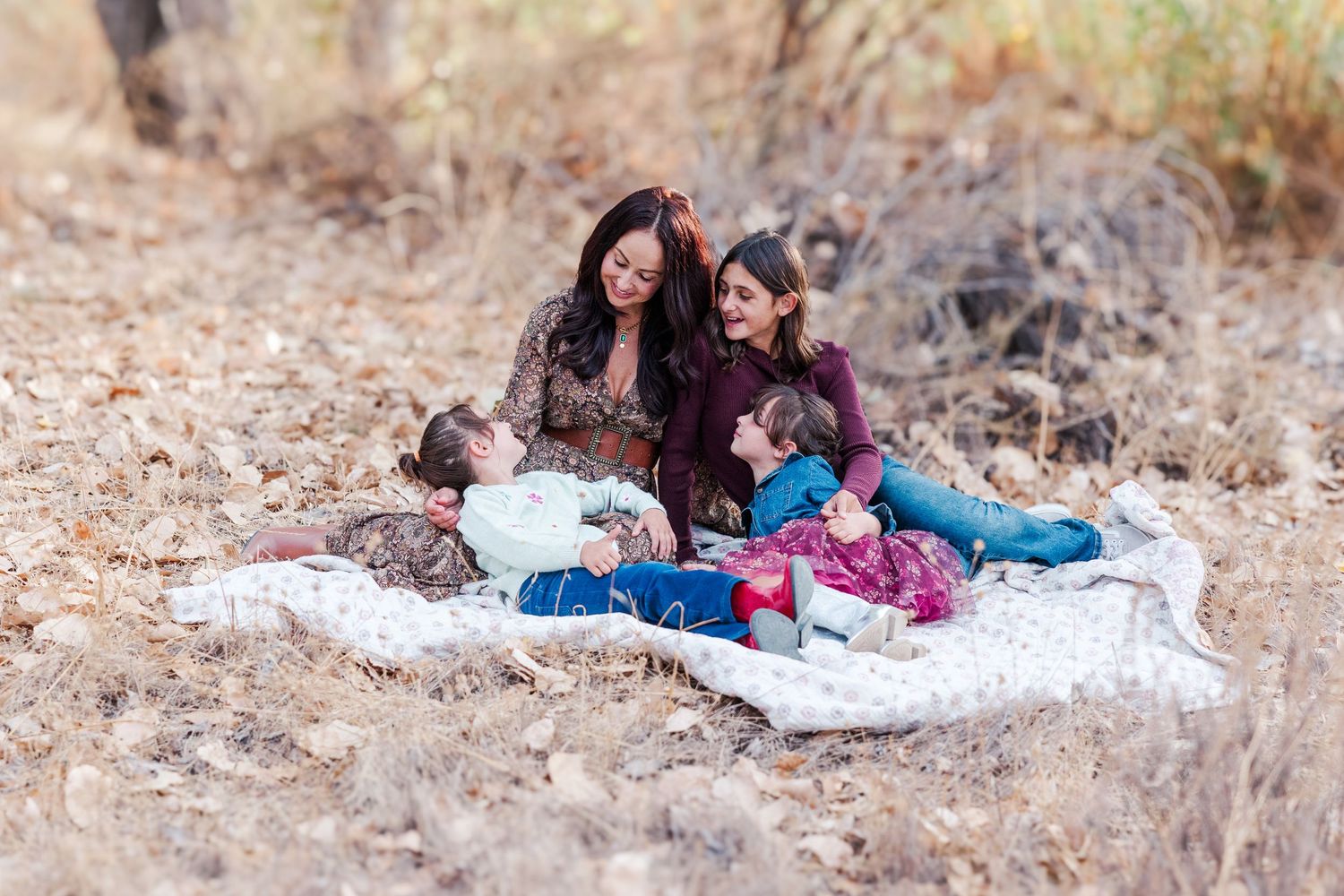 Family relaxing together on a white blanket in a dried grass field on a sunny day.