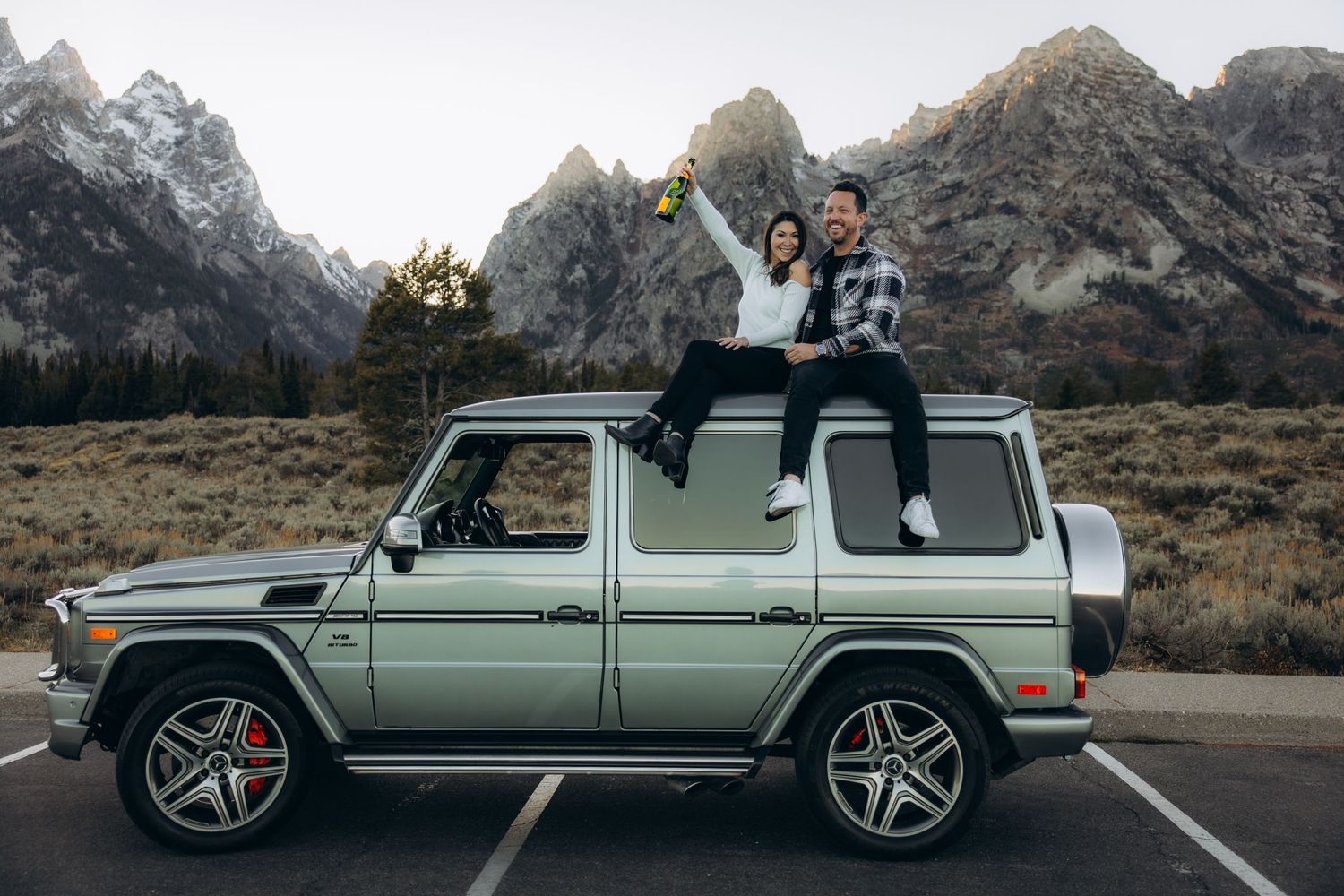 A silver Mercedes G-Wagon SUV parked in front of majestic mountain peaks in a scenic outdoor setting.