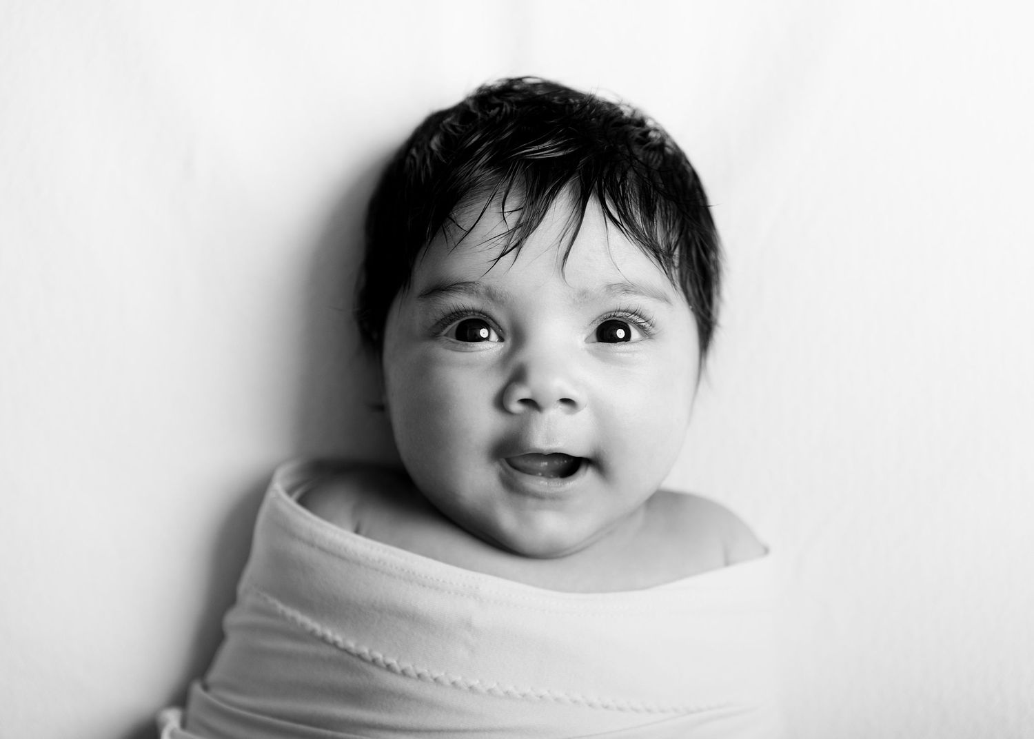 A smiling baby wrapped in white fabric shown in black and white portrait photography against a plain background.