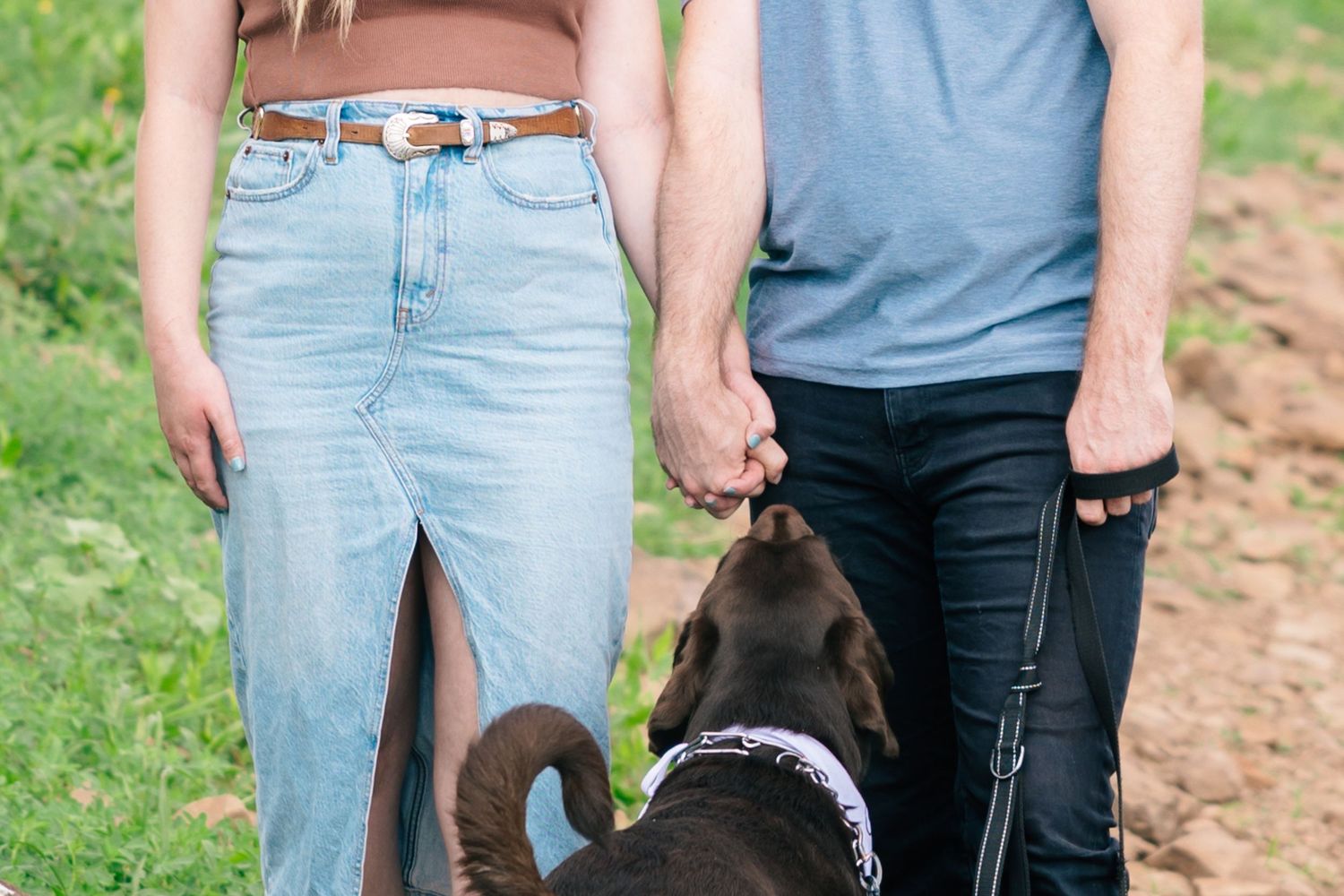 A couple in blue jeans holding hands while walking a black dog on a dirt path outdoors. In Knoxville, TN