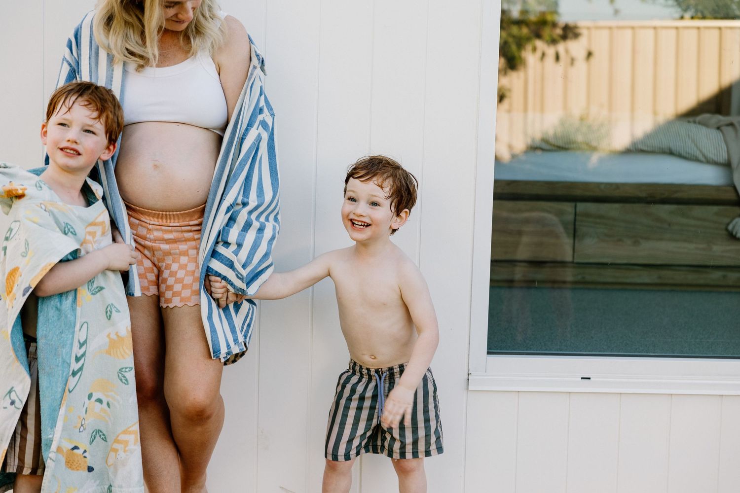 Two young children in swimwear stand beside pregnant woman wearing striped cover up at pool on sunny day.