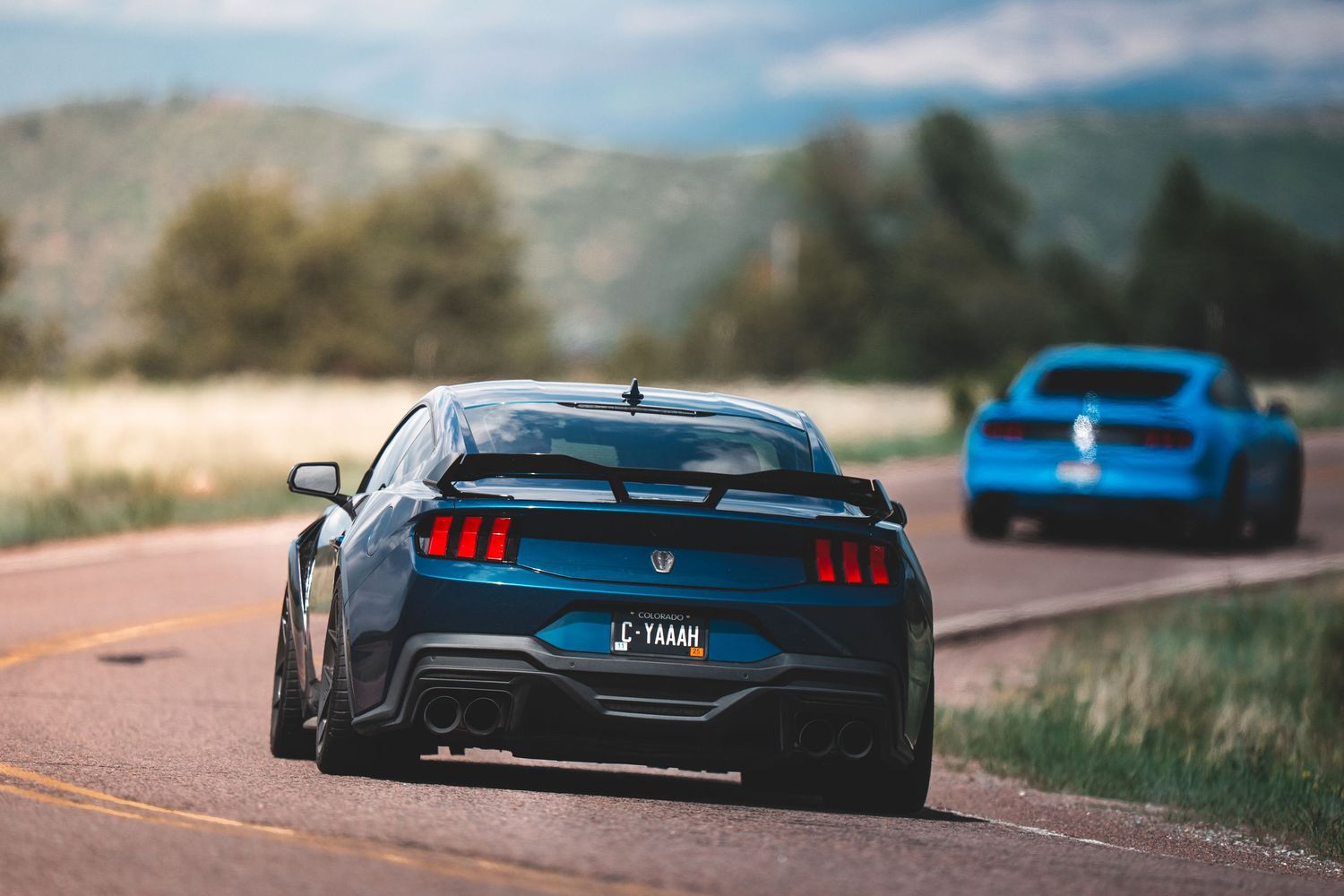 Two Ford Mustangs, one gray and one blue, driving on a winding mountain road with scenic landscape views.