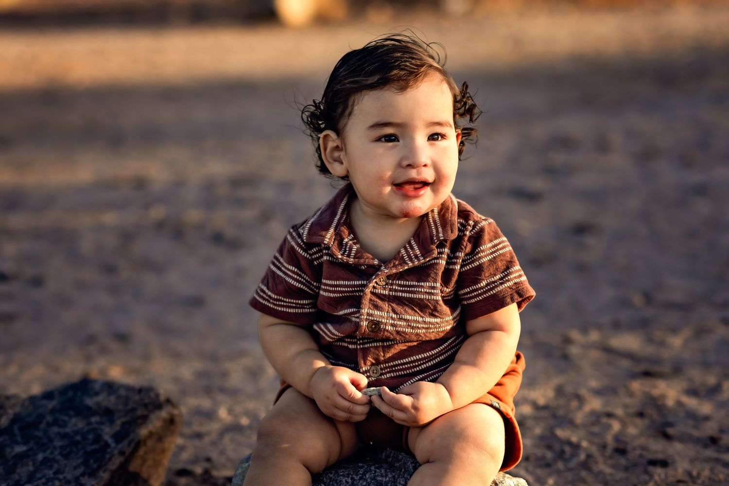 A small child sits and plays in the warm sand during golden hour lighting at sunset on a beach.