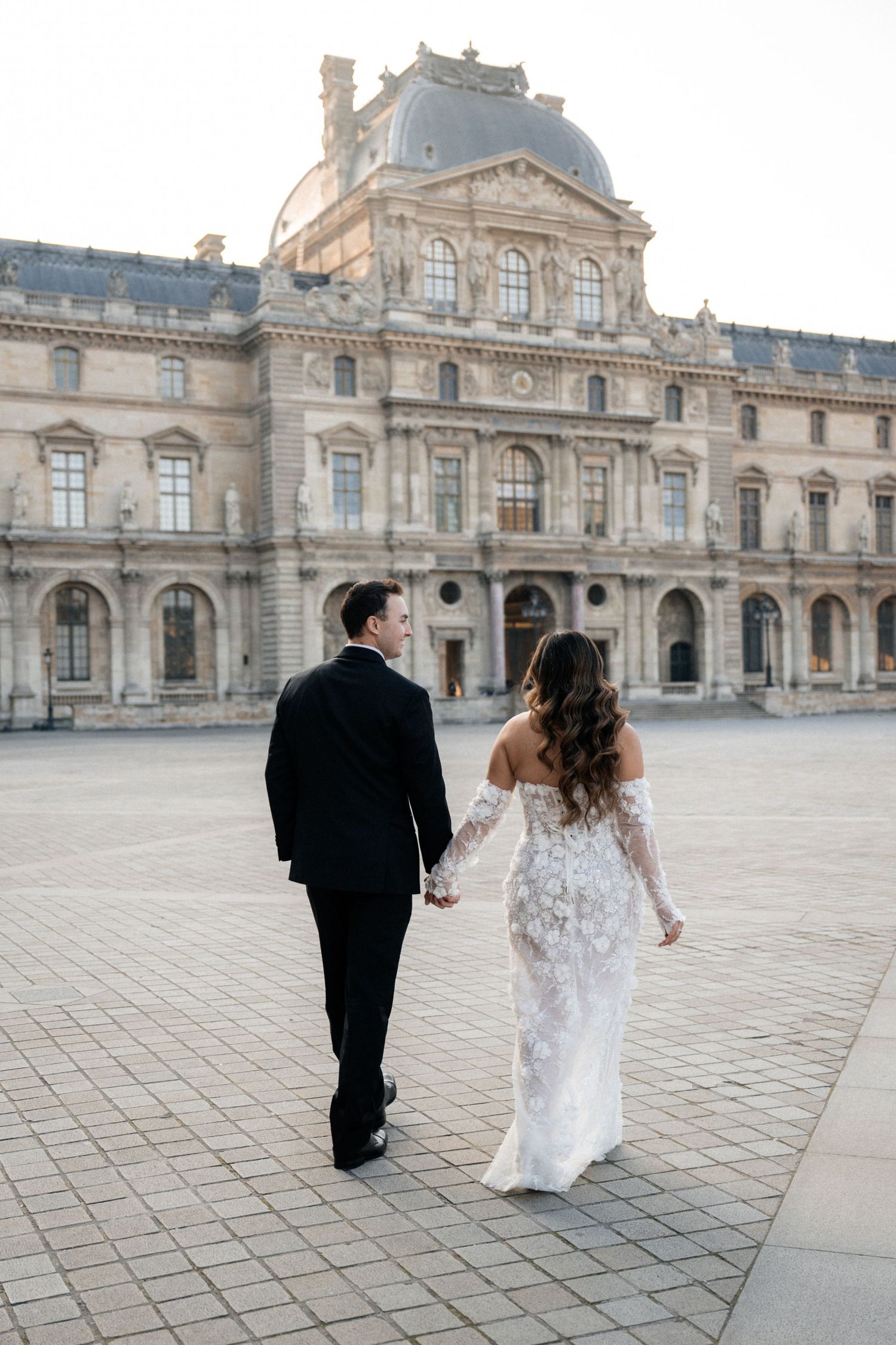 A wedding couple walks in front of the grand Louvre Museum in Paris, with ornate architecture in the background.
