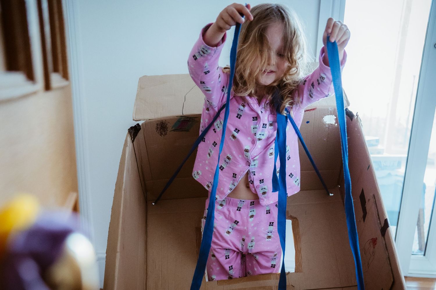 Child in pink pajamas plays with ribbons while standing inside a large cardboard box near a window.