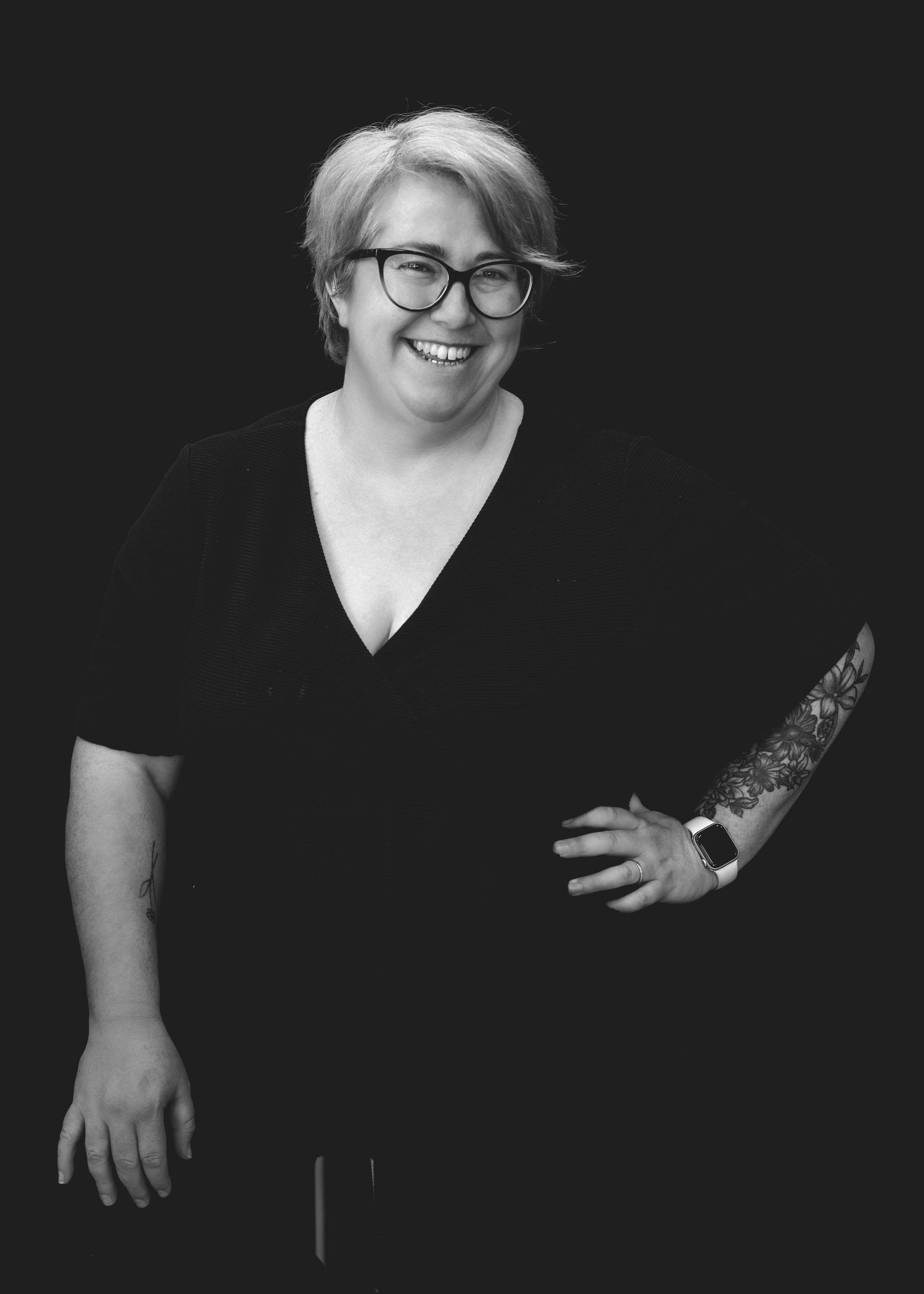 Black and white portrait of someone wearing glasses and a dark v-neck top, smiling brightly at the camera.