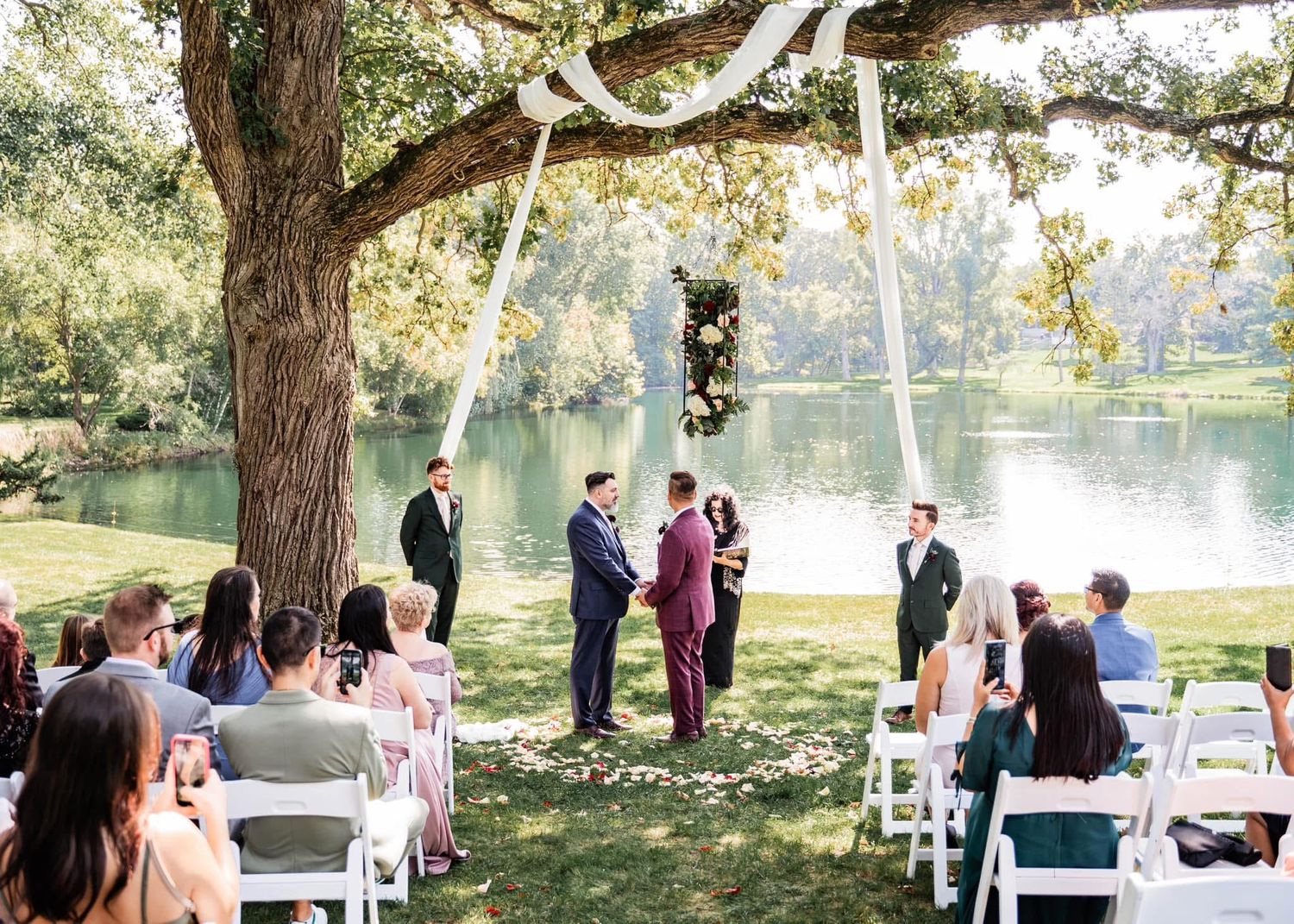 A wedding ceremony takes place outdoors under a large tree by a serene lake with white chairs arranged for guests.